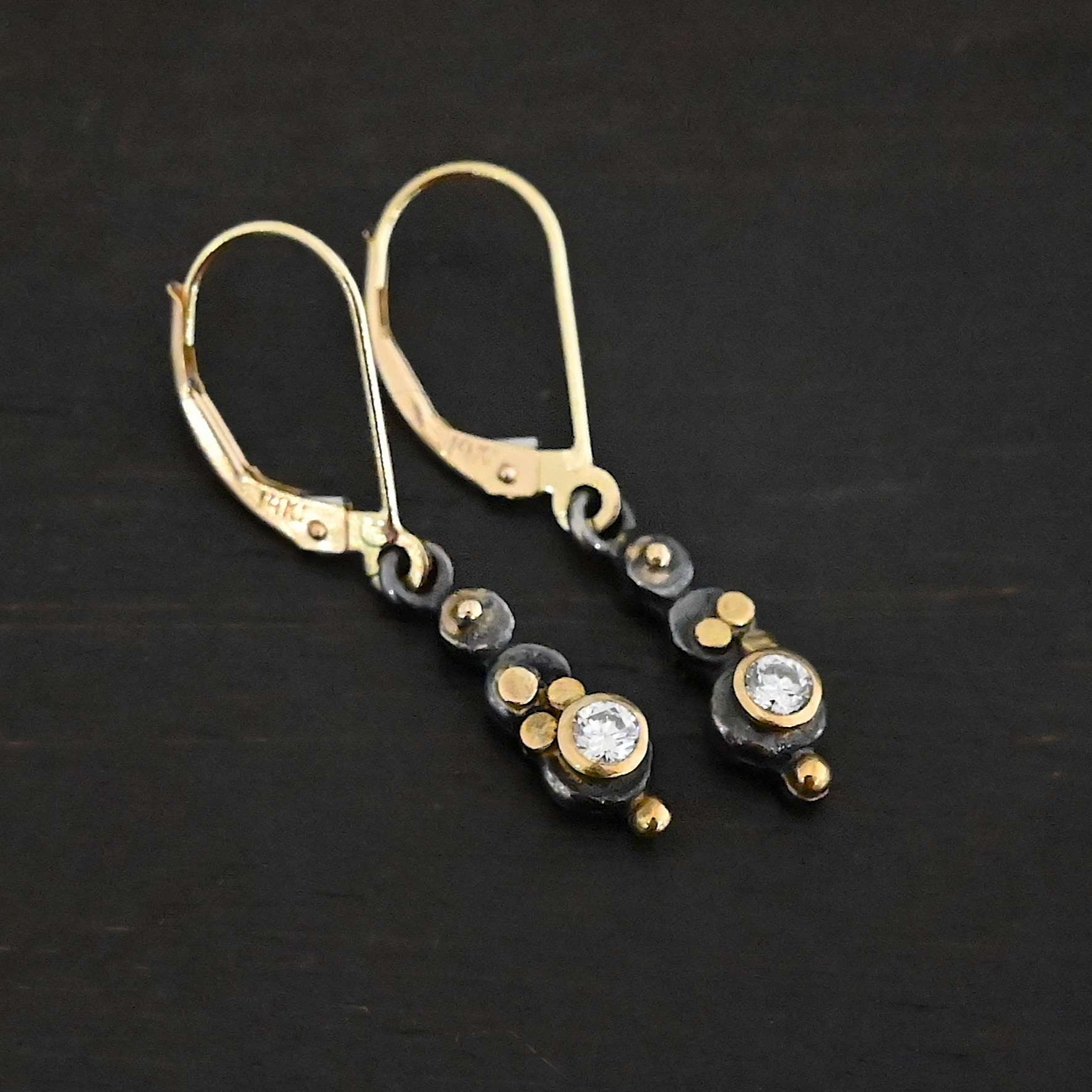 Prairie Princess Earrings Close up of the "Prairie Princess" Earrings. Made out of sterling silver, gold, and diamonds. There are three silver dots that are flat, going from smallest to biggest from the top to the bottom. On the smallest top silver hammered dot is a gold dot that is not hammered. On the second hammered dot are three gold dots that are hammered. On the final hammered dot is a diamond that is set in gold. On the bottom of the earring is a gold dot. Earrings are on gold leverback ear wires. Earrings are laying against a dark background.