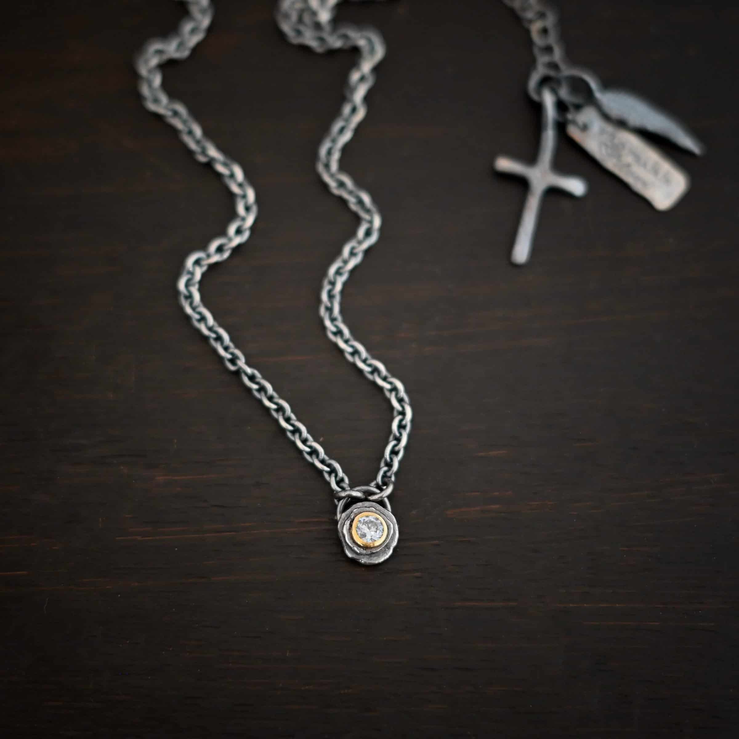 Audrey Hepburn Necklace Photo of the "Audrey Hepburn" Necklace. There is a small diamond in the center of a small circle pendant. The diamond is set in gold. There are two layers of sterling silver that are both hammered flat dots. One layer of silver is smaller, and sits on top of the bigger layer of silver. The gold is on the very top of the pendant. There is a small loop soldered to the back of the necklace in which two silver jump rings hang from. Each end of the chain hangs on the jump rings. Necklace is lying against a dark background.