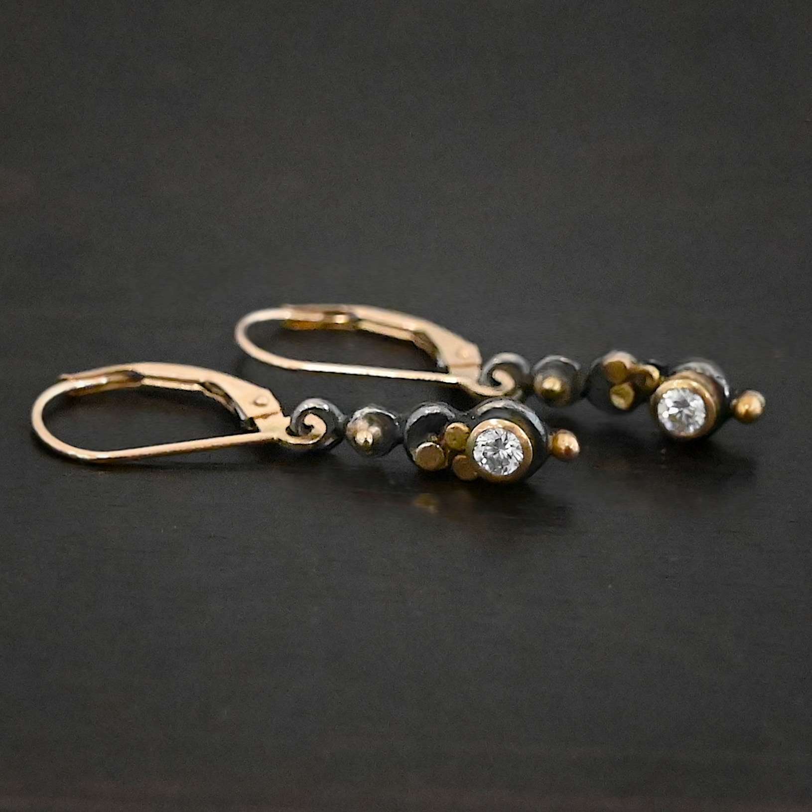 Prairie Princess Earrings Close up of the "Prairie Princess" Earrings. Made out of sterling silver, gold, and diamonds. There are three silver dots that are flat, going from smallest to biggest from the top to the bottom. On the smallest top silver hammered dot is a gold dot that is not hammered. On the second hammered dot are three gold dots that are hammered. On the final hammered dot is a diamond that is set in gold. On the bottom of the earring is a gold dot. Earrings are on gold leverback ear wires. Earrings are laying against a dark background.