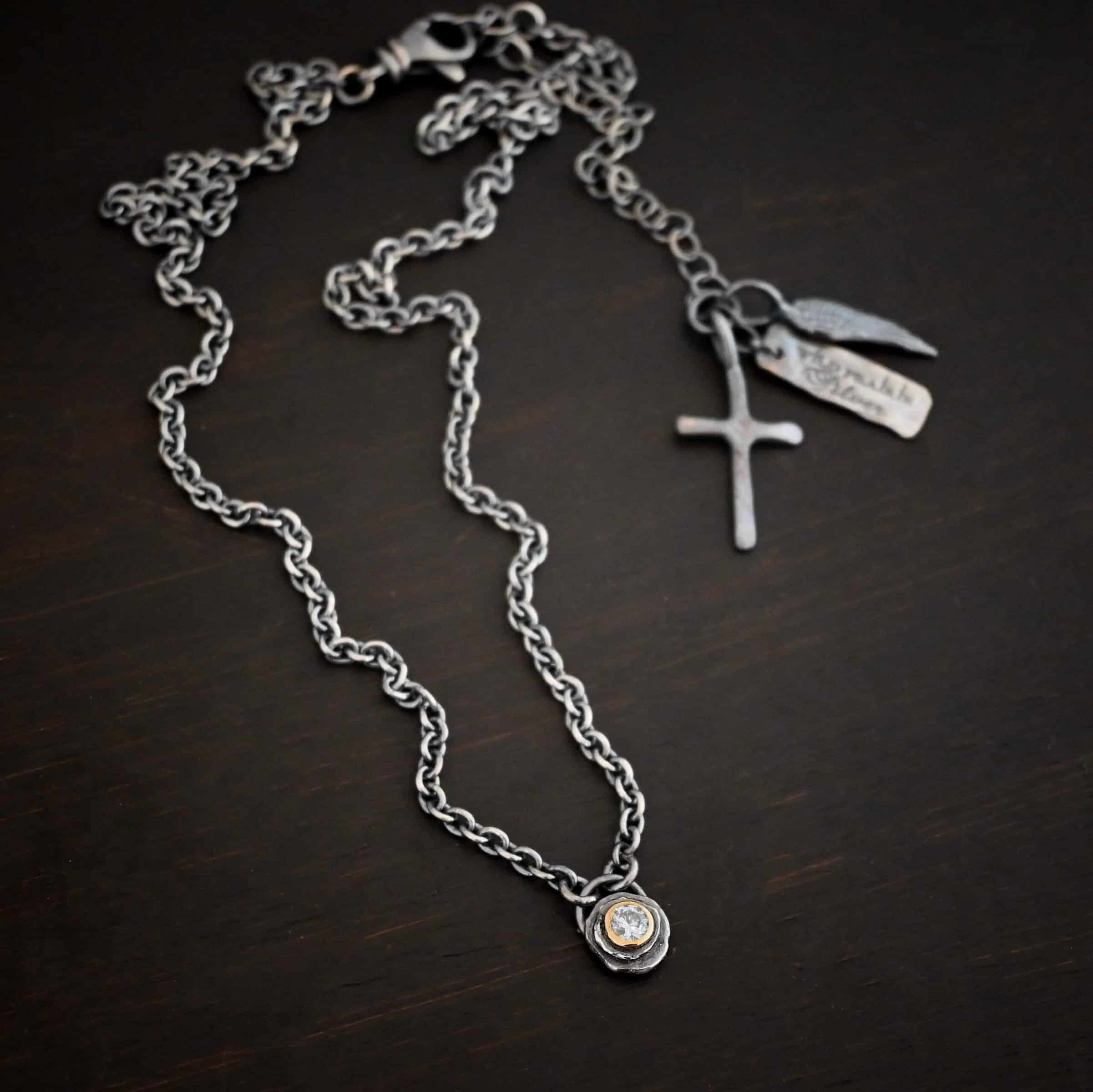 Audrey Hepburn Necklace Photo of the "Audrey Hepburn" Necklace. There is a small diamond in the center of a small circle pendant. The diamond is set in gold. There are two layers of sterling silver that are both hammered flat dots. One layer of silver is smaller, and sits on top of the bigger layer of silver. The gold is on the very top of the pendant. There is a small loop soldered to the back of the necklace in which two silver jump rings hang from. Each end of the chain hangs on the jump rings. Necklace is lying against a dark background.