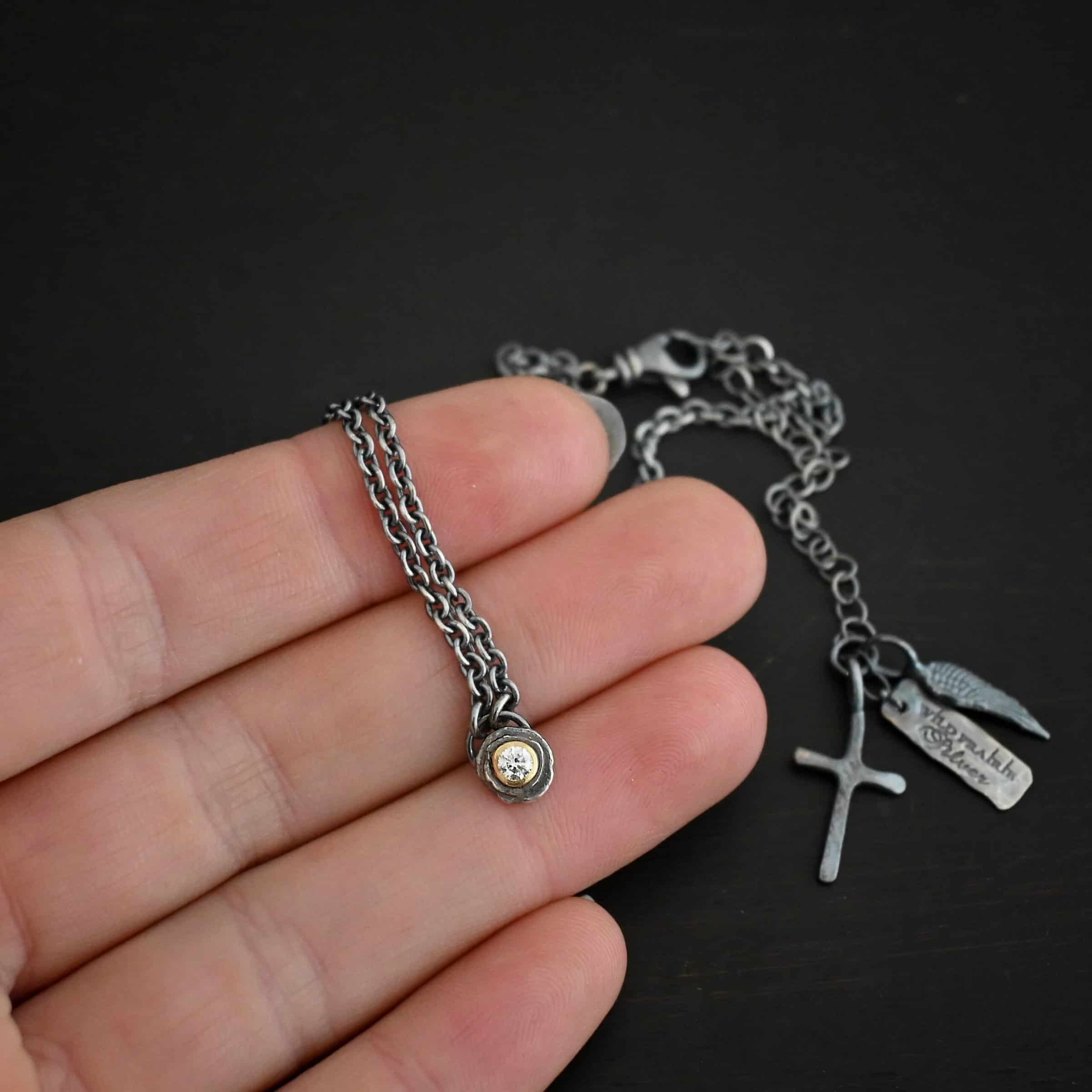 Audrey Hepburn Necklace Photo of the "Audrey Hepburn" Necklace. There is a small diamond in the center of a small circle pendant. The diamond is set in gold. There are two layers of sterling silver that are both hammered flat dots. One layer of silver is smaller, and sits on top of the bigger layer of silver. The gold is on the very top of the pendant. There is a small loop soldered to the back of the necklace in which two silver jump rings hang from. Each end of the chain hangs on the jump rings. Necklace is lying against the fingertips of a hand to demonstrate size.