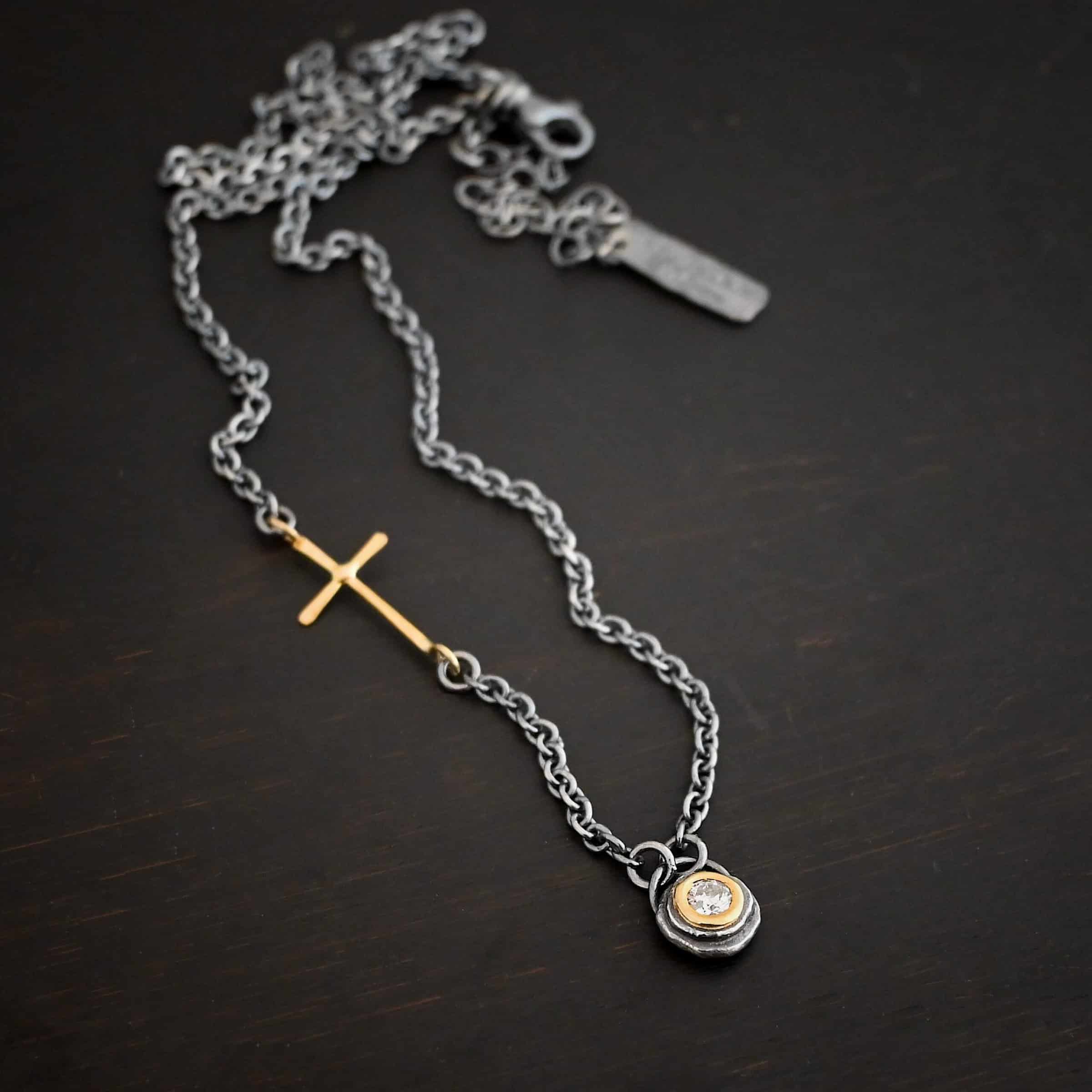 Devotion Necklace Photo of the "Devotion" Necklace. There is a small diamond in the center of a small circle pendant. The diamond is set in gold. There are two layers of sterling silver that are both hammered flat dots. One layer of silver is smaller, and sits on top of the bigger layer of silver. The gold is on the very top of the pendant. There is a small loop soldered to the back of the necklace in which two silver jump rings hang from. Each end of the chain hangs on the jump rings. There is a large gold cross that is attached on the left side of the chain that is attached to the chain with two jump rings. The end and top of the cross have a loop. The bottom loop attaches to a jump ring on the chain, as well as the top loop, so the cross is fully connected to the chain. Necklace is lying against a dark background.