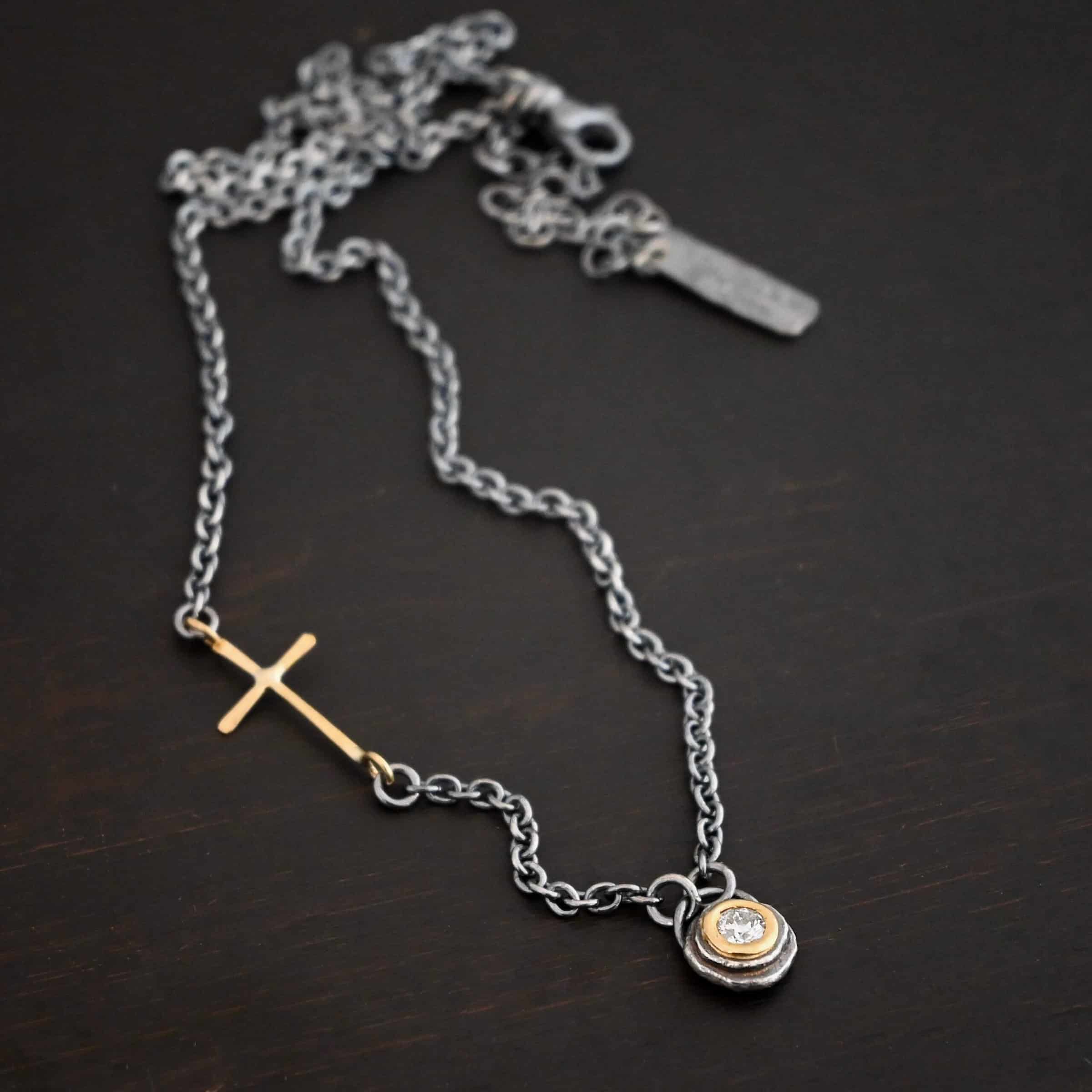 Devotion Necklace Photo of the "Devotion" Necklace. There is a small diamond in the center of a small circle pendant. The diamond is set in gold. There are two layers of sterling silver that are both hammered flat dots. One layer of silver is smaller, and sits on top of the bigger layer of silver. The gold is on the very top of the pendant. There is a small loop soldered to the back of the necklace in which two silver jump rings hang from. Each end of the chain hangs on the jump rings. There is a large gold cross that is attached on the left side of the chain that is attached to the chain with two jump rings. The end and top of the cross have a loop. The bottom loop attaches to a jump ring on the chain, as well as the top loop, so the cross is fully connected to the chain. Necklace is lying against a dark background.