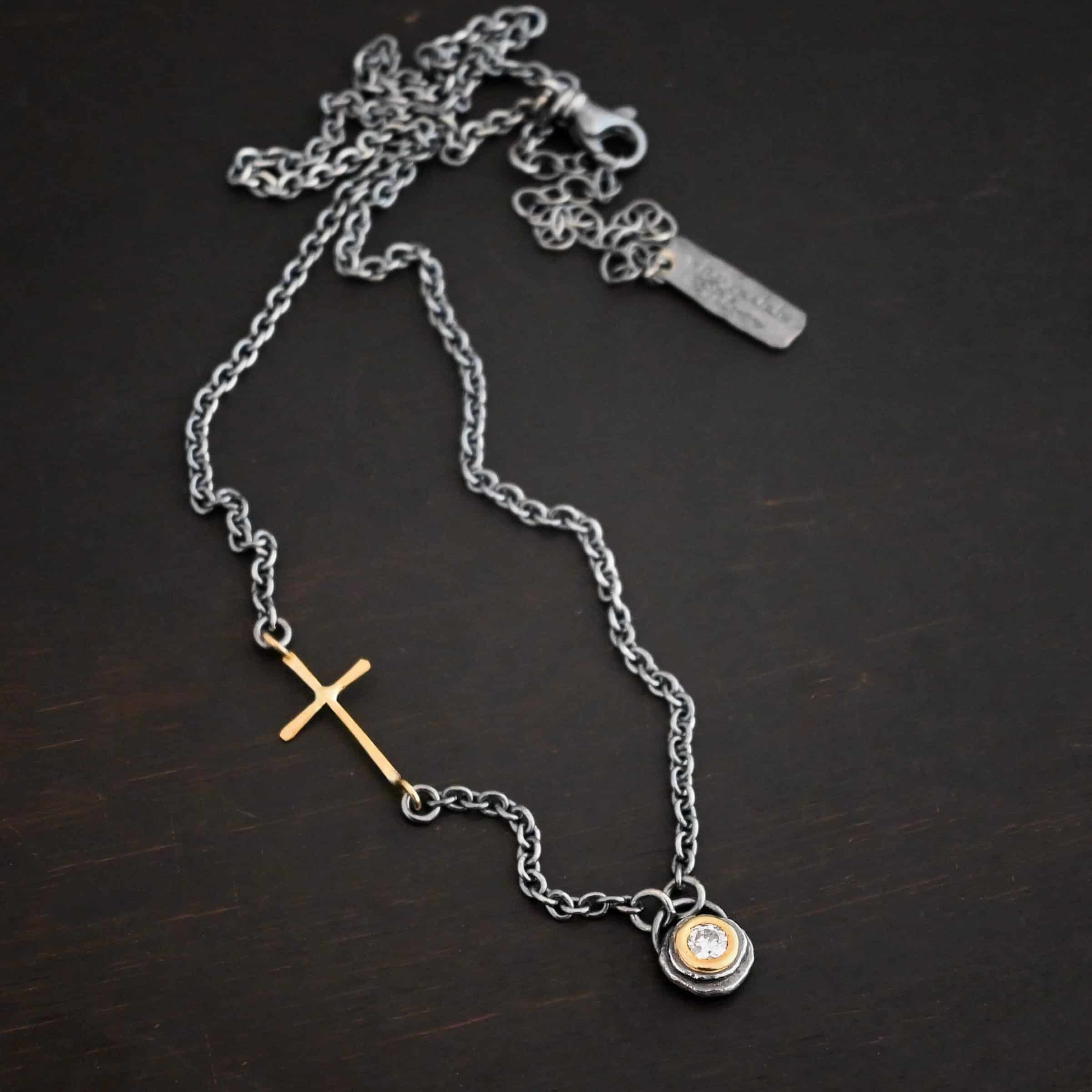 Devotion Necklace Photo of the "Devotion" Necklace. There is a small diamond in the center of a small circle pendant. The diamond is set in gold. There are two layers of sterling silver that are both hammered flat dots. One layer of silver is smaller, and sits on top of the bigger layer of silver. The gold is on the very top of the pendant. There is a small loop soldered to the back of the necklace in which two silver jump rings hang from. Each end of the chain hangs on the jump rings. There is a large gold cross that is attached on the left side of the chain that is attached to the chain with two jump rings. The end and top of the cross have a loop. The bottom loop attaches to a jump ring on the chain, as well as the top loop, so the cross is fully connected to the chain. Necklace is lying against a dark background.