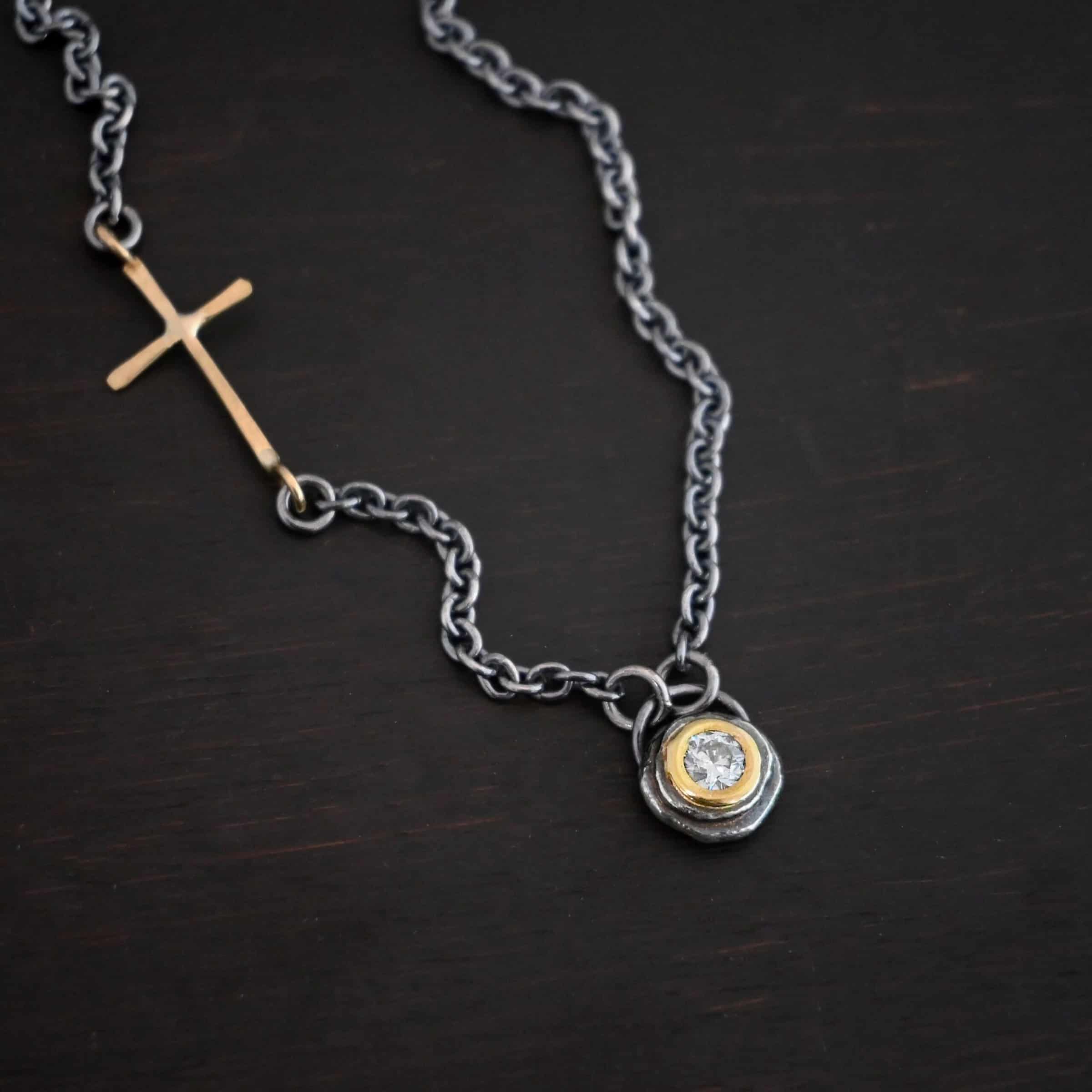 Devotion Necklace Photo of the "Devotion" Necklace. There is a small diamond in the center of a small circle pendant. The diamond is set in gold. There are two layers of sterling silver that are both hammered flat dots. One layer of silver is smaller, and sits on top of the bigger layer of silver. The gold is on the very top of the pendant. There is a small loop soldered to the back of the necklace in which two silver jump rings hang from. Each end of the chain hangs on the jump rings. There is a large gold cross that is attached on the left side of the chain that is attached to the chain with two jump rings. The end and top of the cross have a loop. The bottom loop attaches to a jump ring on the chain, as well as the top loop, so the cross is fully connected to the chain. Necklace is lying against a dark background.