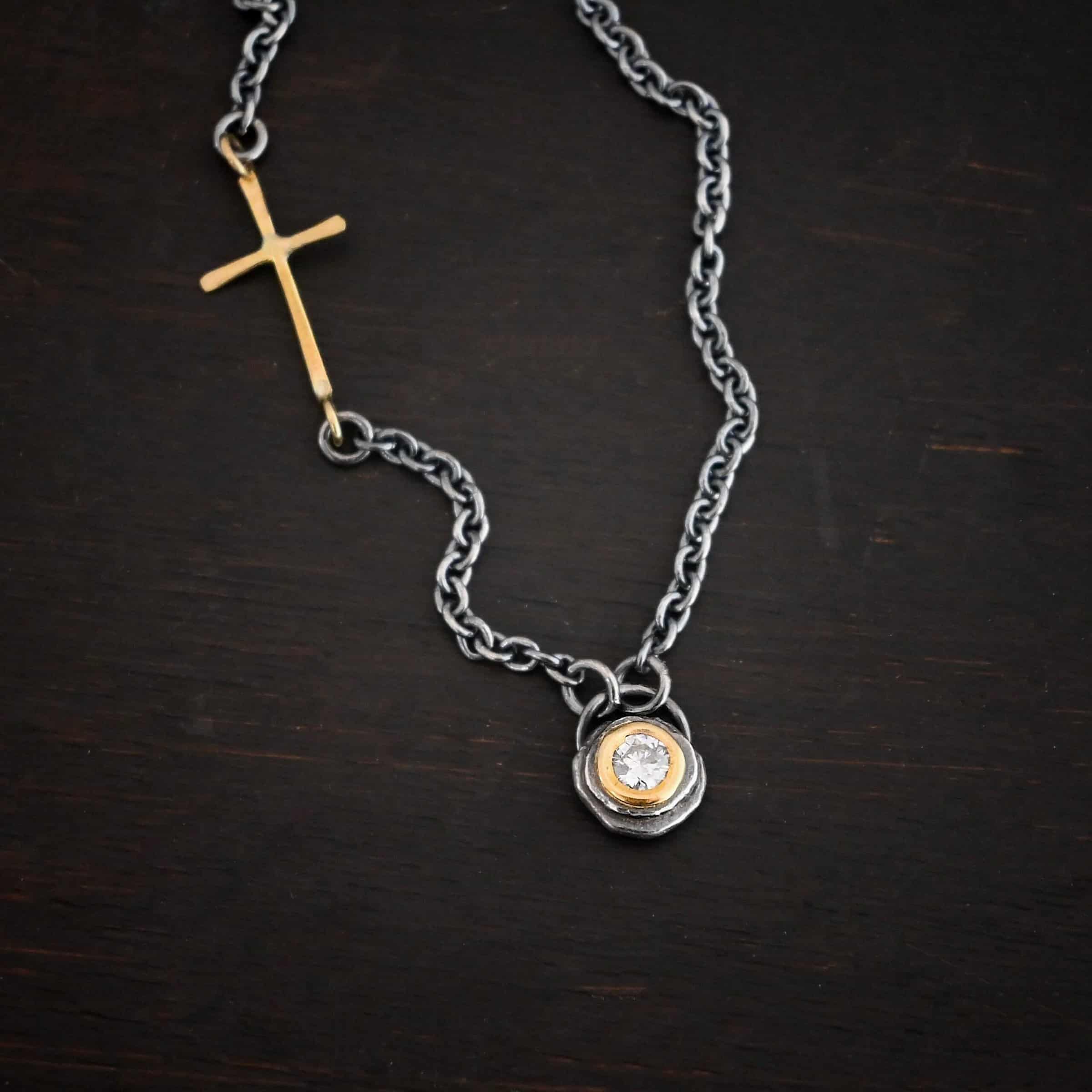 Devotion Necklace Photo of the "Devotion" Necklace. There is a small diamond in the center of a small circle pendant. The diamond is set in gold. There are two layers of sterling silver that are both hammered flat dots. One layer of silver is smaller, and sits on top of the bigger layer of silver. The gold is on the very top of the pendant. There is a small loop soldered to the back of the necklace in which two silver jump rings hang from. Each end of the chain hangs on the jump rings. There is a large gold cross that is attached on the left side of the chain that is attached to the chain with two jump rings. The end and top of the cross have a loop. The bottom loop attaches to a jump ring on the chain, as well as the top loop, so the cross is fully connected to the chain. Necklace is lying against a dark background.