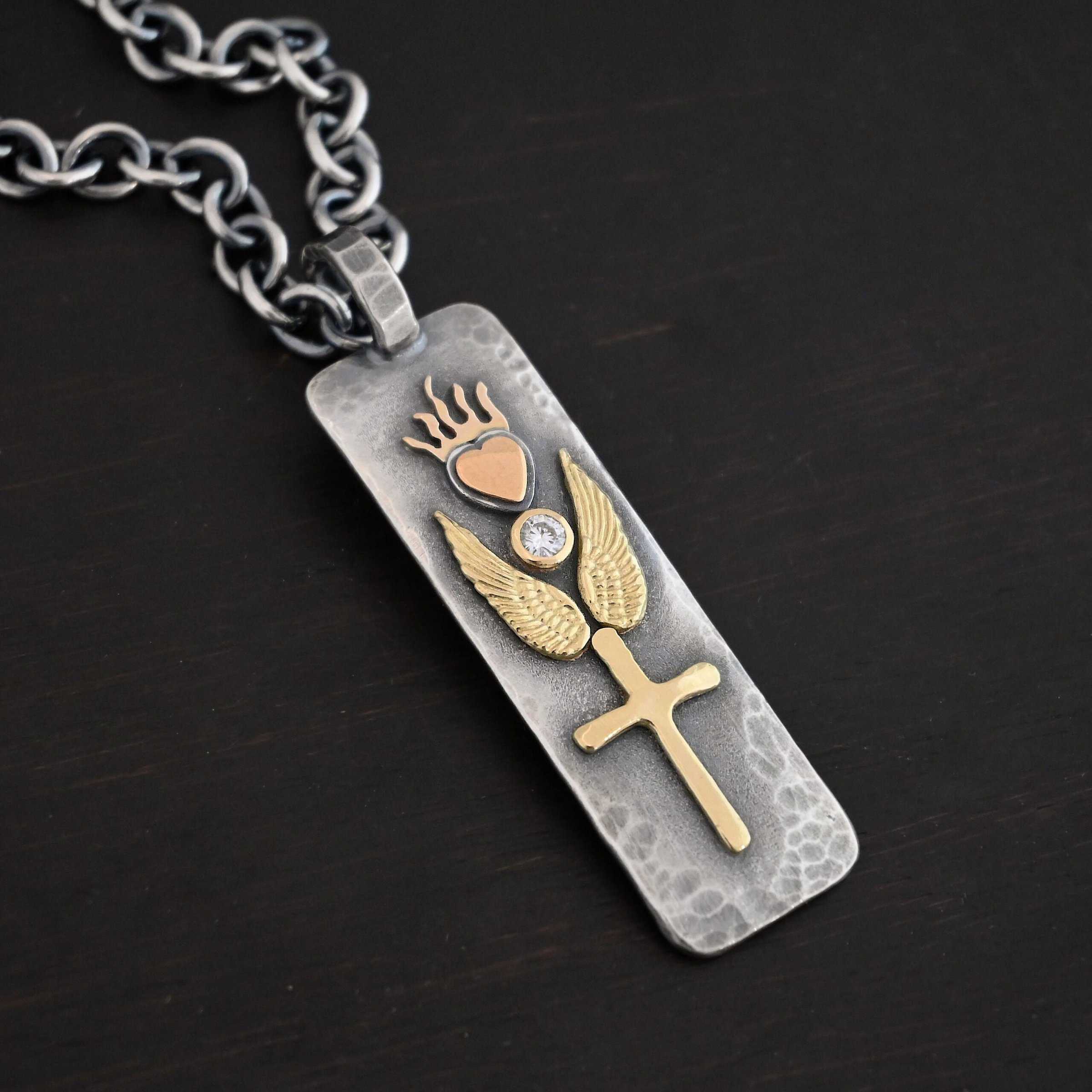 Protect Necklace Photo of the "Protect" Necklace. Necklace is made out of silver and gold. The pendant is a rectangle shaped silver tag that hangs vertically. It is large. On the top of the rectangle is a gold heart with gold flames dancing across the top of it. On the bottom, beneath the heart is a diamond that is set in gold. On each side of the diamond is a gold angel wing. Beneath the gold angel wings in the center of them and on the bottom of the rectangle pendant is a gold cross. There is a thick jump ring soldered to the top of the pendant that the chain of the necklace goes through. Necklace is laying against a dark background.