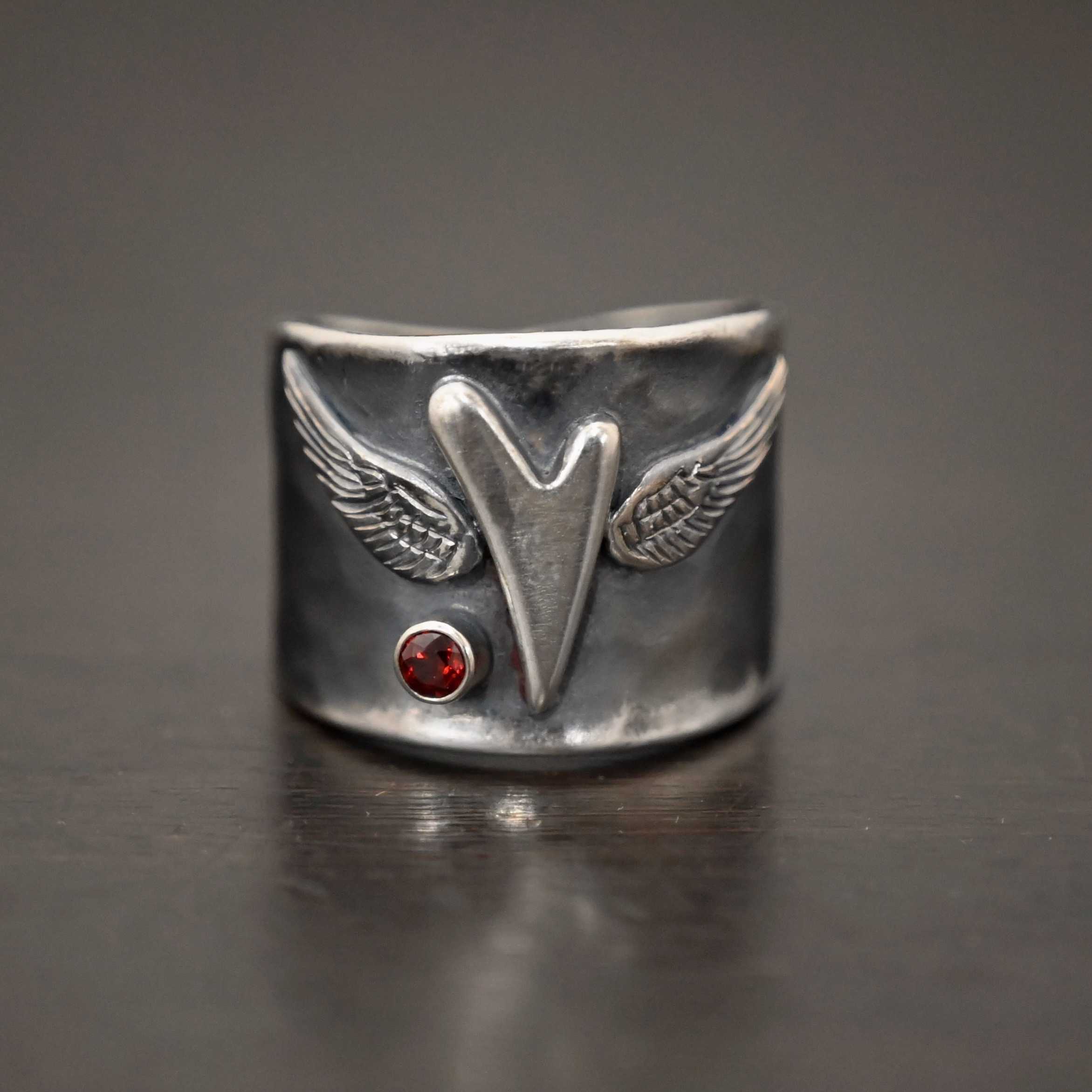 Close up of the "Wild Prairie" Ring. Made out of sterling silver and garnet. There is a silver heart in the center of the ring. It is a tall heart that stretches from almost the top of the ring to the bottom. There are sterling silver angel wings sprouting from each side of the heart. There is a garnet set in silver on the left side of the ring underneath one of the angel wings. Ring is sitting against a dark background.