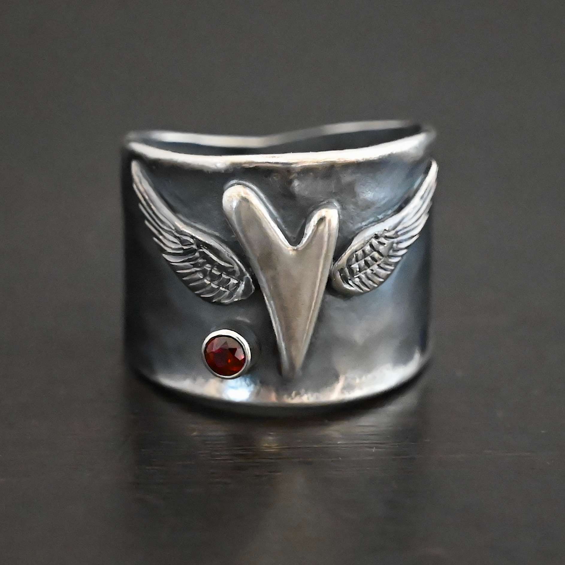 Close up of the "Wild Prairie" Ring. Made out of sterling silver and garnet. There is a silver heart in the center of the ring. It is a tall heart that stretches from almost the top of the ring to the bottom. There are sterling silver angel wings sprouting from each side of the heart. There is a garnet set in silver on the left side of the ring underneath one of the angel wings. Ring is sitting against a dark background.