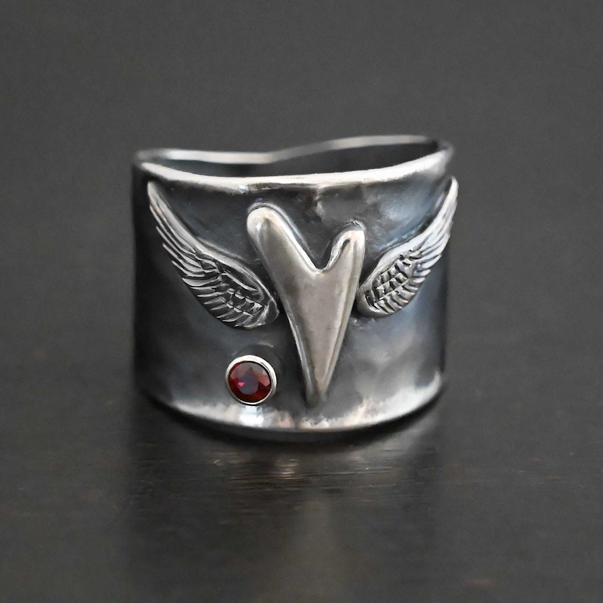 Close up of the "Wild Prairie" Ring. Made out of sterling silver and garnet. There is a silver heart in the center of the ring. It is a tall heart that stretches from almost the top of the ring to the bottom. There are sterling silver angel wings sprouting from each side of the heart. There is a garnet set in silver on the left side of the ring underneath one of the angel wings. Ring is sitting against a dark background.