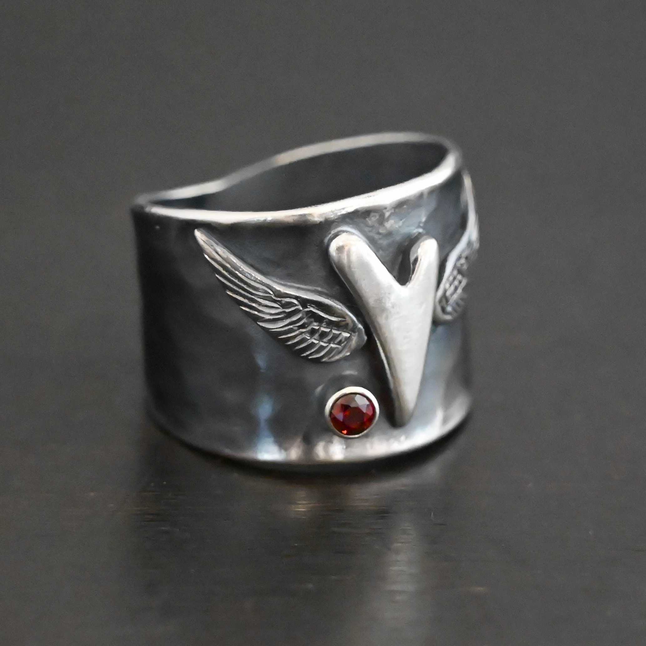 Close up of the "Wild Prairie" Ring. Made out of sterling silver and garnet. There is a silver heart in the center of the ring. It is a tall heart that stretches from almost the top of the ring to the bottom. There are sterling silver angel wings sprouting from each side of the heart. There is a garnet set in silver on the left side of the ring underneath one of the angel wings. Ring is sitting against a dark background.