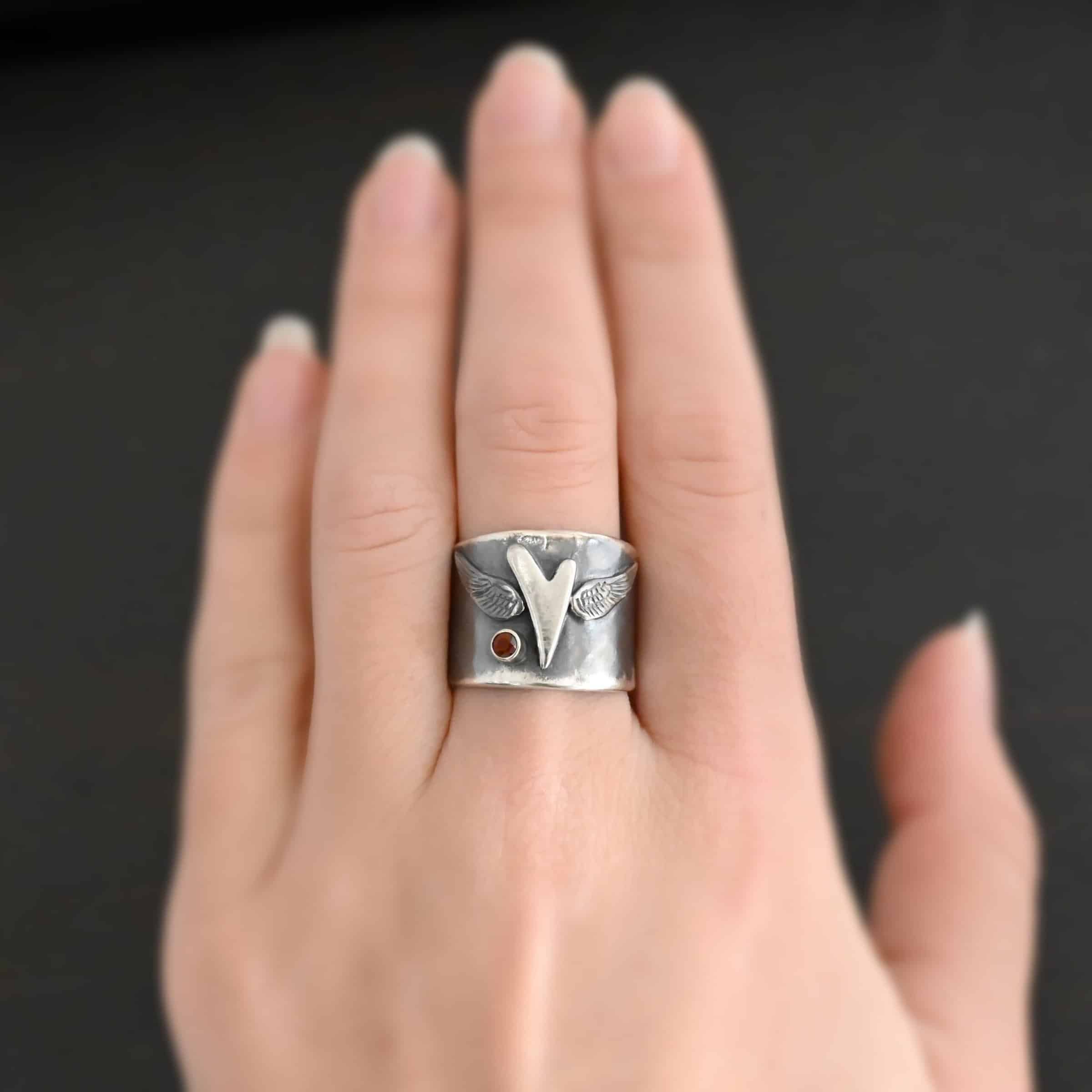Close up of the "Wild Prairie" Ring. Made out of sterling silver and garnet. There is a silver heart in the center of the ring. It is a tall heart that stretches from almost the top of the ring to the bottom. There are sterling silver angel wings sprouting from each side of the heart. There is a garnet set in silver on the left side of the ring underneath one of the angel wings. Ring is worn on a models middle finger to demonstrate size.