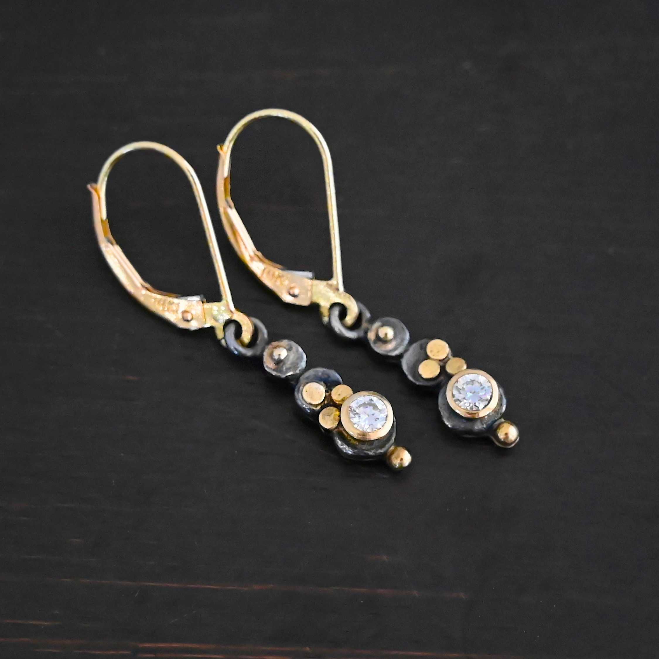 Prairie Princess Earrings Close up of the "Prairie Princess" Earrings. Made out of sterling silver, gold, and diamonds. There are three silver dots that are flat, going from smallest to biggest from the top to the bottom. On the smallest top silver hammered dot is a gold dot that is not hammered. On the second hammered dot are three gold dots that are hammered. On the final hammered dot is a diamond that is set in gold. On the bottom of the earring is a gold dot. Earrings are on gold leverback ear wires. Earrings are laying against a dark background.