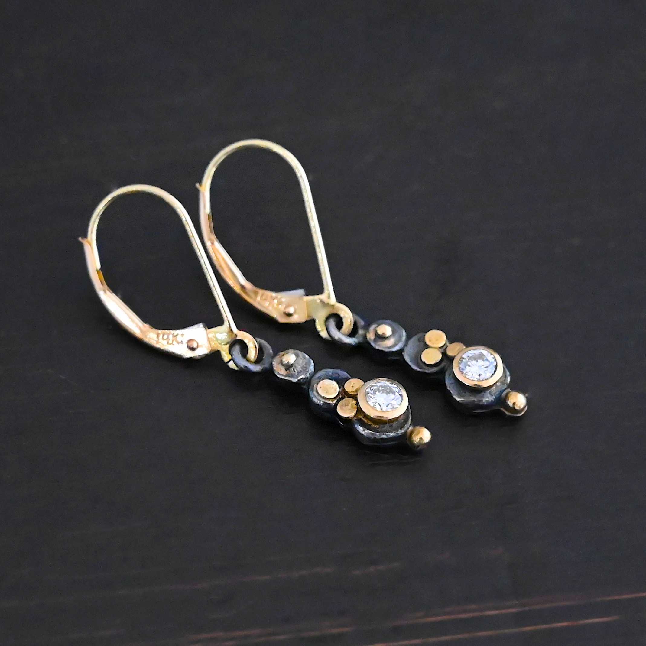Prairie Princess Earrings Close up of the "Prairie Princess" Earrings. Made out of sterling silver, gold, and diamonds. There are three silver dots that are flat, going from smallest to biggest from the top to the bottom. On the smallest top silver hammered dot is a gold dot that is not hammered. On the second hammered dot are three gold dots that are hammered. On the final hammered dot is a diamond that is set in gold. On the bottom of the earring is a gold dot. Earrings are on gold leverback ear wires. Earrings are laying against a dark background.