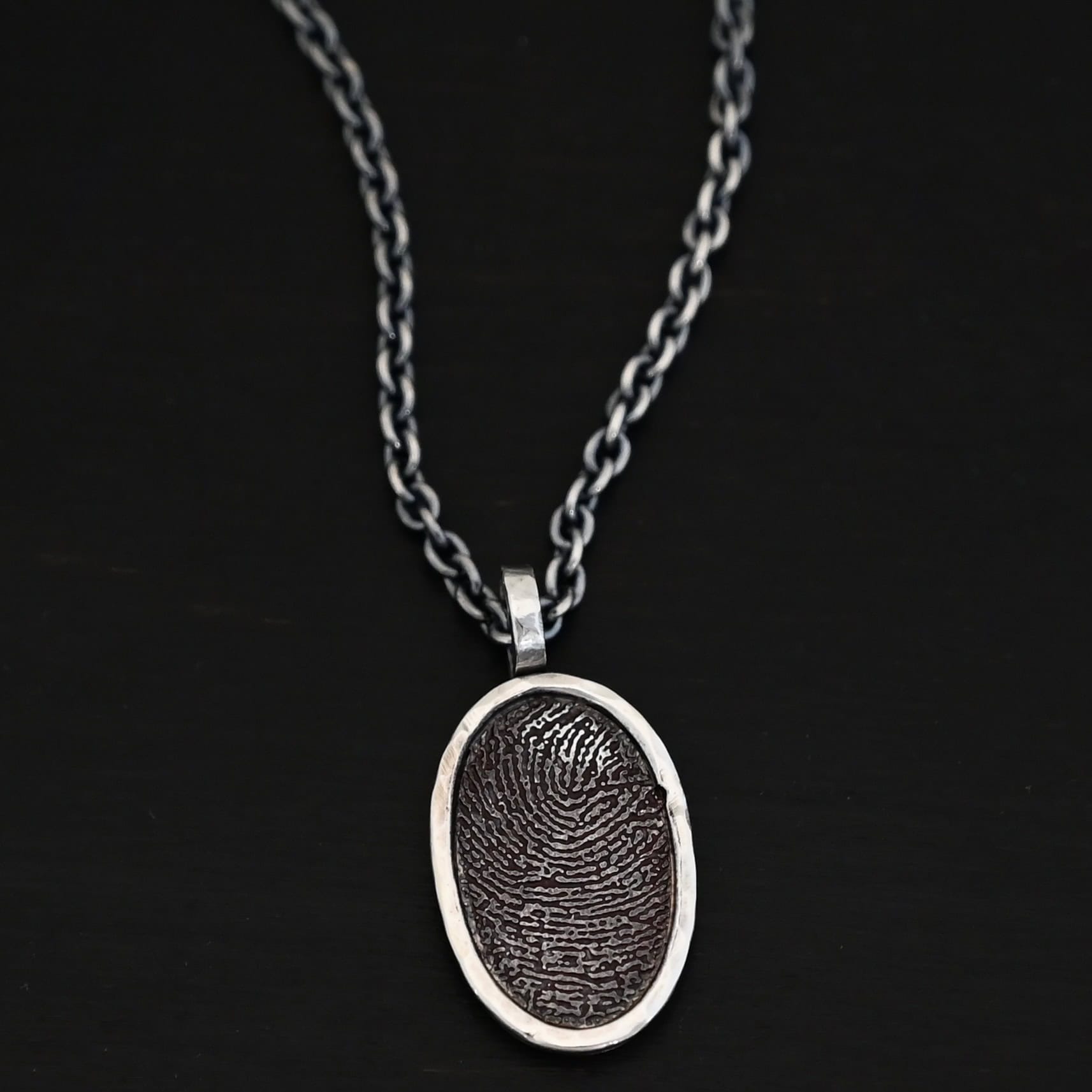 Photo of the "Holding You Always" Necklace. Made out of sterling silver. Necklace has an engraved fingerprint in the center for the pendant. The fingerprint is filled and colored in with black to emphasize the fingerprint. Going around the fingerprint is more sterling silver in an oval shape to cover the outside of the edges of the pendant. There is a thick silver stationary loop soldered on the top of the necklace to connect it to the chain. Necklace is laying against a dark background.