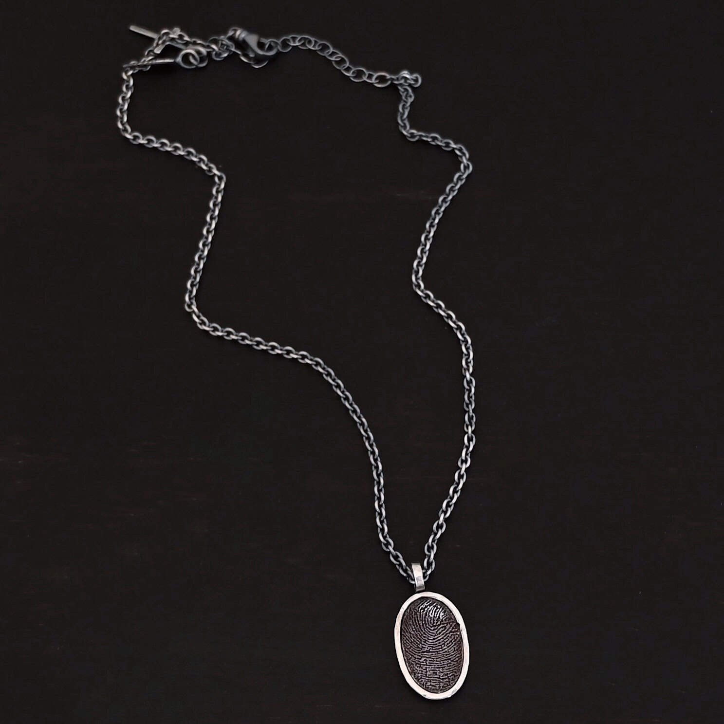 Photo of the "Holding You Always" Necklace. Made out of sterling silver. Necklace has an engraved fingerprint in the center for the pendant. The fingerprint is filled and colored in with black to emphasize the fingerprint. Going around the fingerprint is more sterling silver in an oval shape to cover the outside of the edges of the pendant. There is a thick silver stationary loop soldered on the top of the necklace to connect it to the chain. Necklace is laying against a dark background.