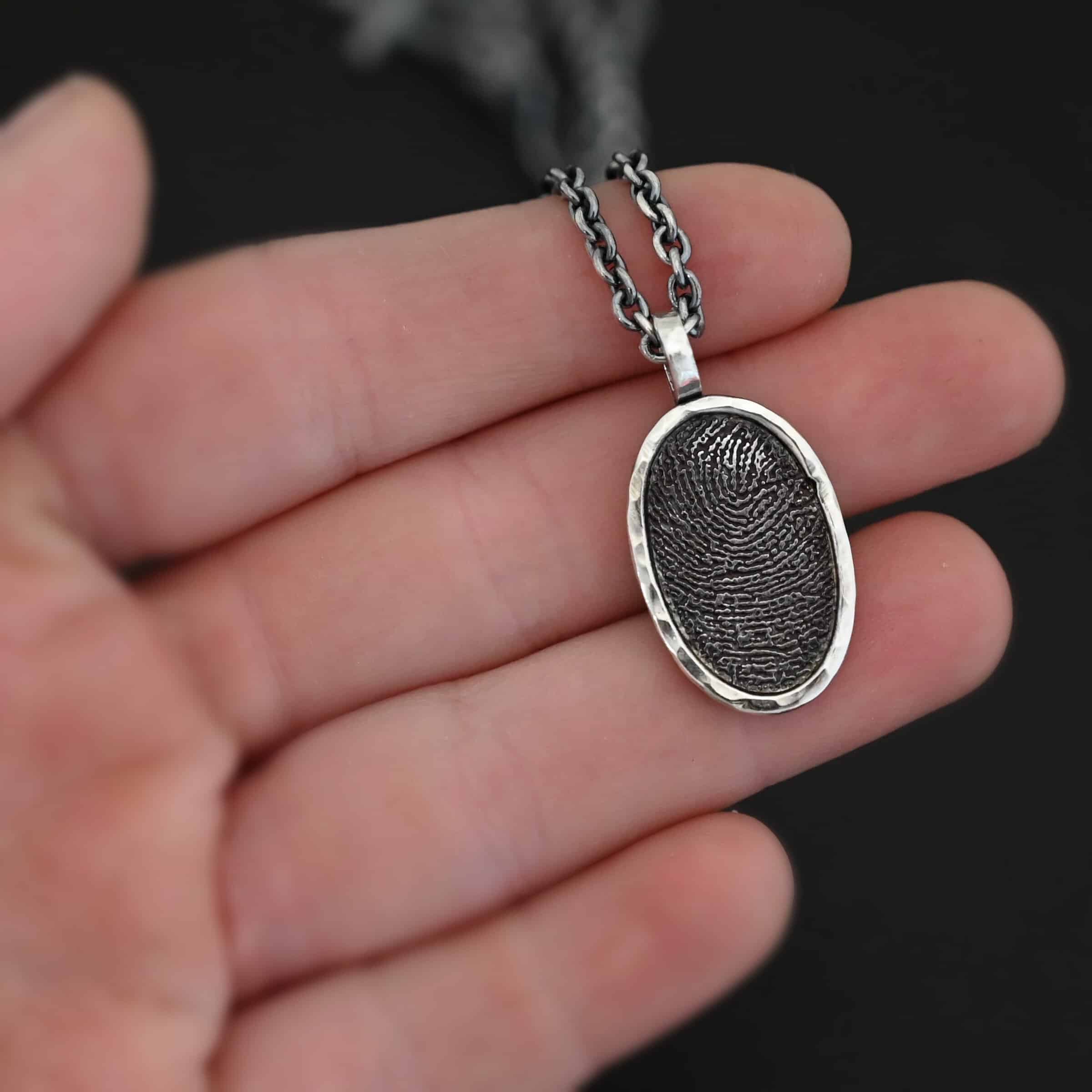 Photo of the "Holding You Always" Necklace. Made out of sterling silver. Necklace has an engraved fingerprint in the center for the pendant. The fingerprint is filled and colored in with black to emphasize the fingerprint. Going around the fingerprint is more sterling silver in an oval shape to cover the outside of the edges of the pendant. There is a thick silver stationary loop soldered on the top of the necklace to connect it to the chain. Necklace is laying on the fingertips of a model to demonstrate size of the pendant.
