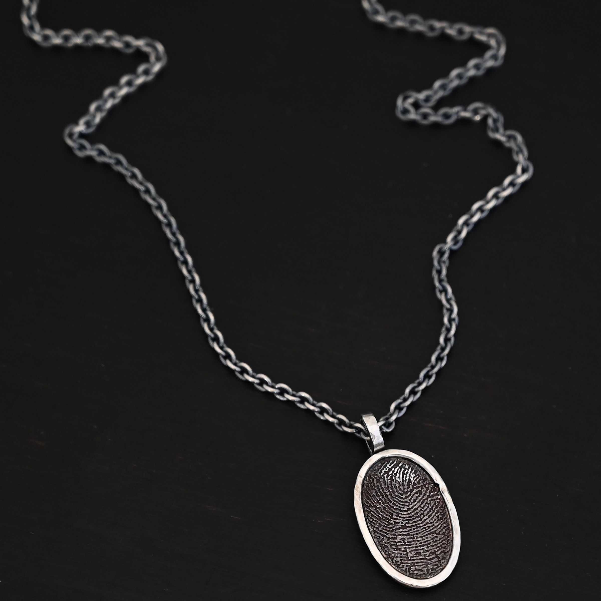 Photo of the "Holding You Always" Necklace. Made out of sterling silver. Necklace has an engraved fingerprint in the center for the pendant. The fingerprint is filled and colored in with black to emphasize the fingerprint. Going around the fingerprint is more sterling silver in an oval shape to cover the outside of the edges of the pendant. There is a thick silver stationary loop soldered on the top of the necklace to connect it to the chain. Necklace is laying against a dark background.