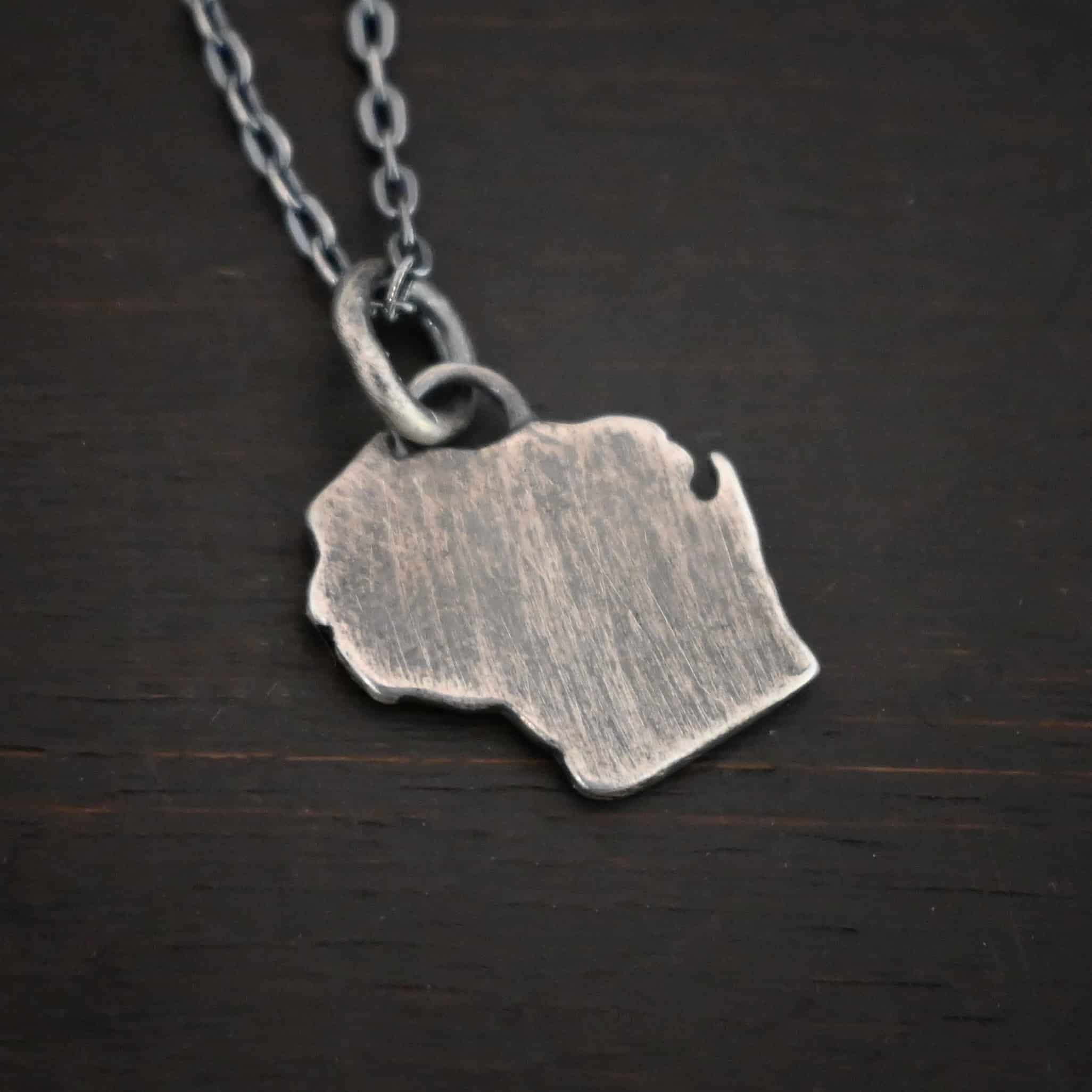 On Wisconsin ox smooth Photo of the "On Wisconsin" Pendant. There is a small, sterling silver Wisconsin charm with a small loop on the top and a jump ring going through it. The charm is oxidized and smooth. Charm is lying against a dark background.