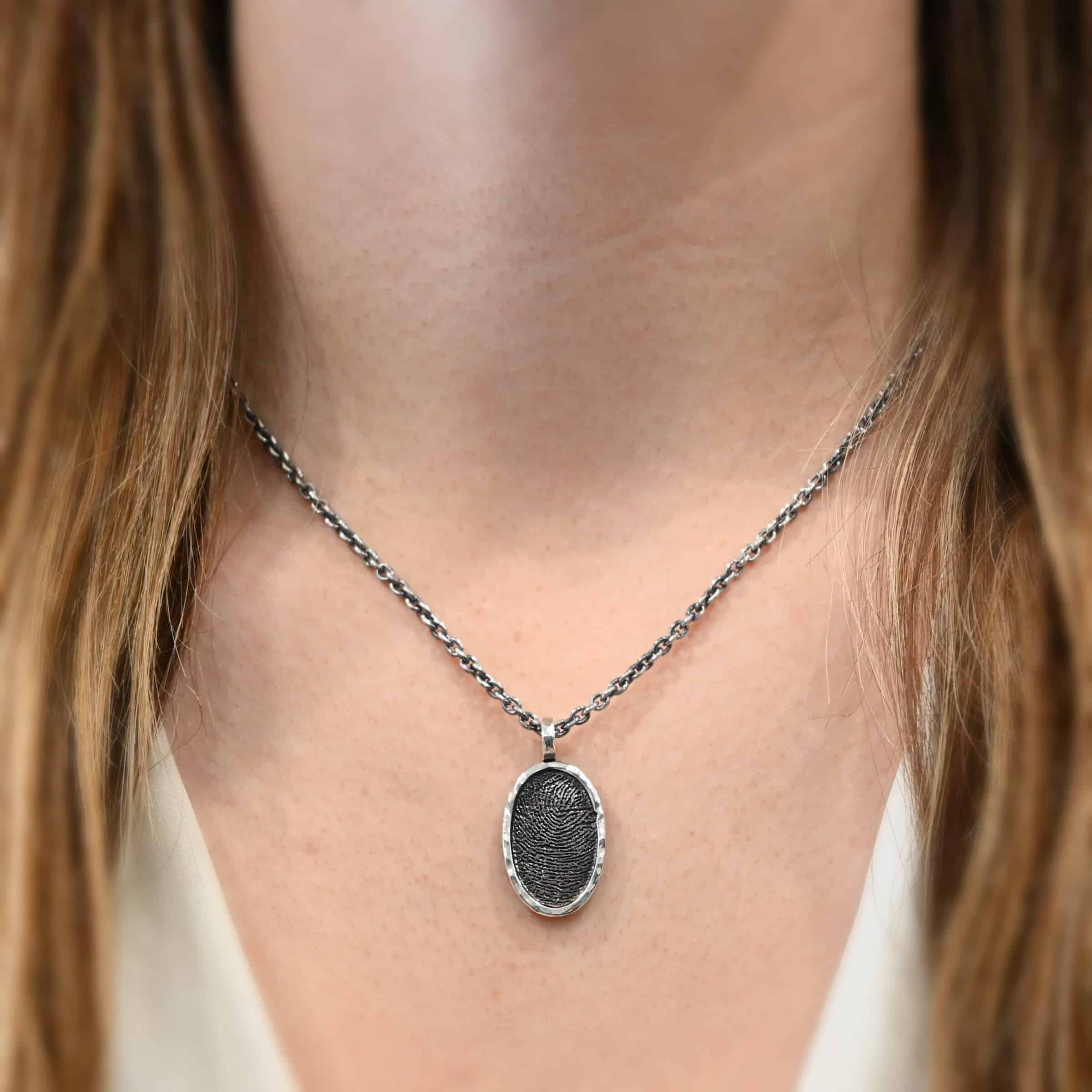 Photo of the "Holding You Always" Necklace. Made out of sterling silver. Necklace has an engraved fingerprint in the center for the pendant. The fingerprint is filled and colored in with black to emphasize the fingerprint. Going around the fingerprint is more sterling silver in an oval shape to cover the outside of the edges of the pendant. There is a thick silver stationary loop soldered on the top of the necklace to connect it to the chain. Necklace is worn by a model at 18 inches long to demonstrate size.