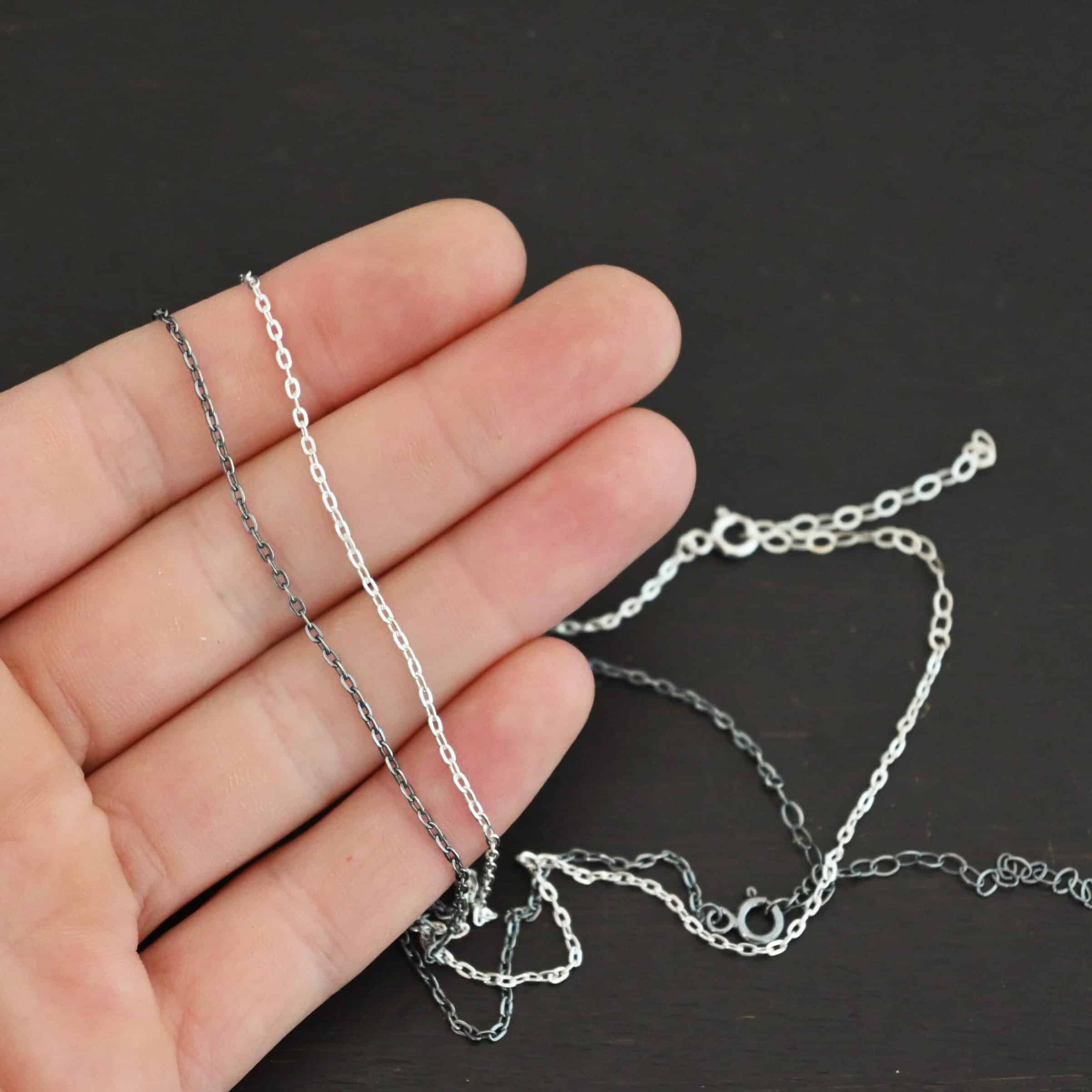 Cassandra Chain "Cassandra" Chain as shown lying against the fingertips of a hand that are lying against a dark background. Chain is shown as shiny and oxidized. Chain has 2x2mm links.