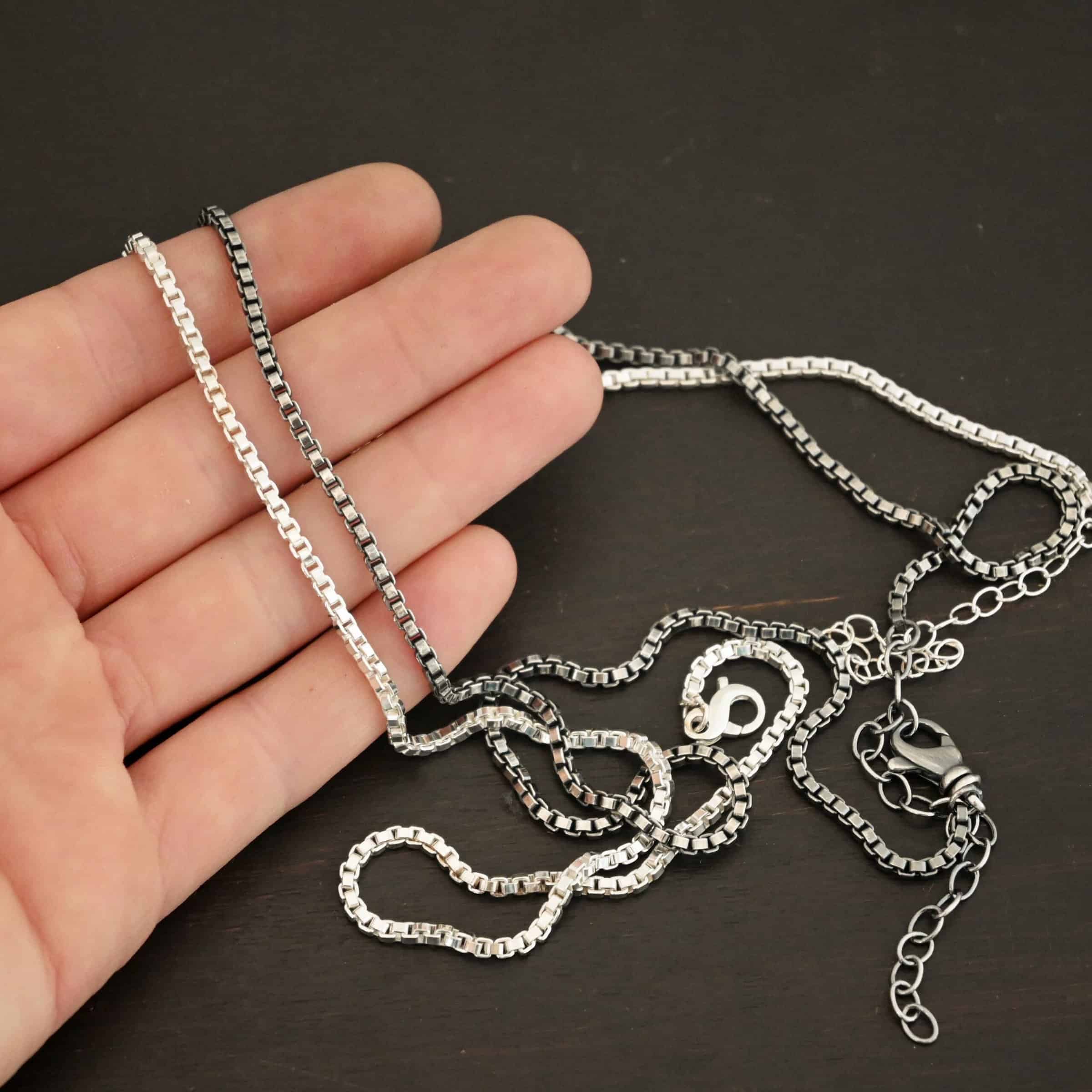 Close up of the "Briggs Box" Chain as both shiny and oxidized. Chain is made out of sterling silver and has box links that make up the chain. The chains are set up against the fingertips of a hand. Heavy Box Chain 2.5mmx1.4mm.