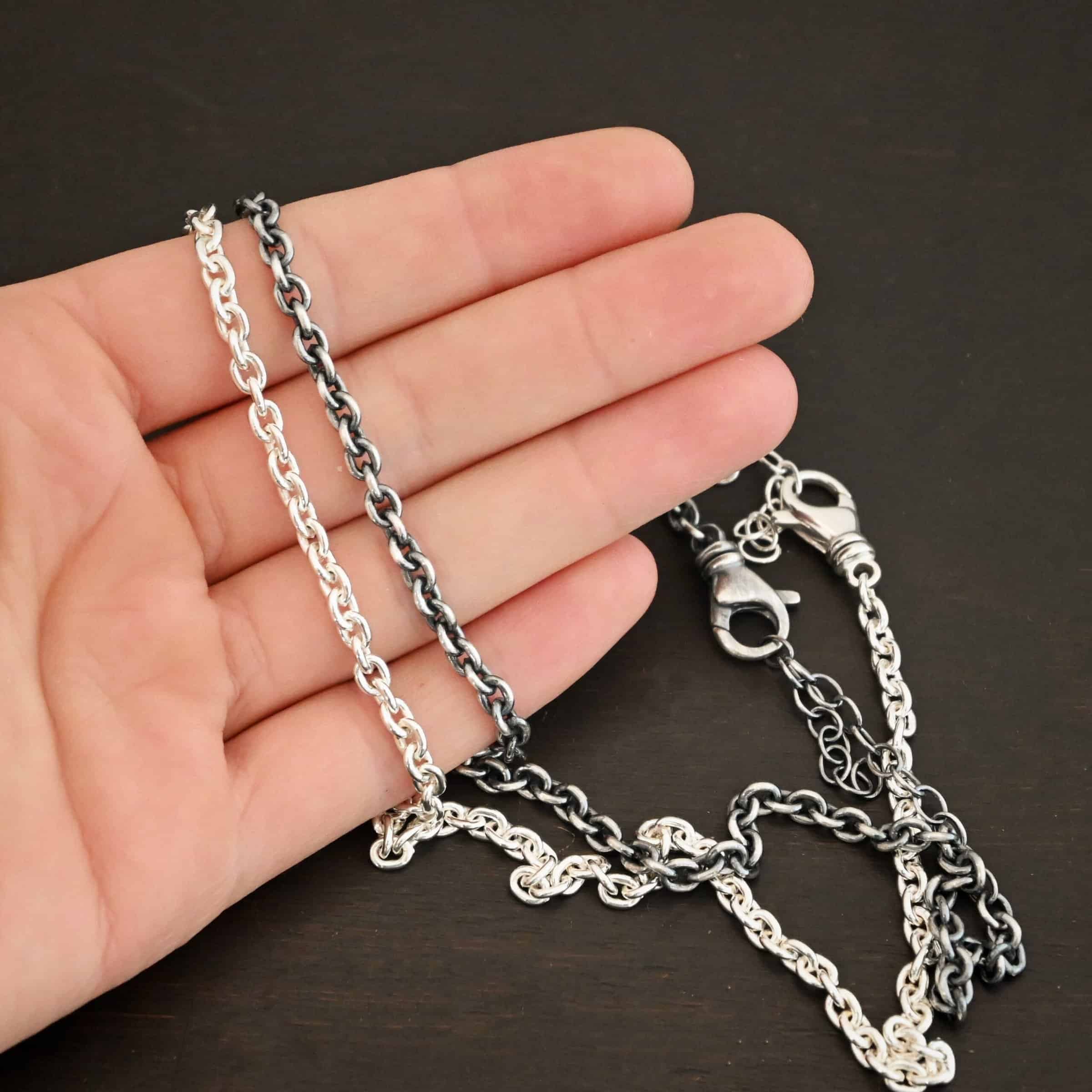 Joan Chain Close up of the "Joan" Chain as both shiny and oxidized. Made out of sterling silver and has thick oval links that measure 6x5mmx1.2mm thick. Chains are set against the fingertips of a hand.