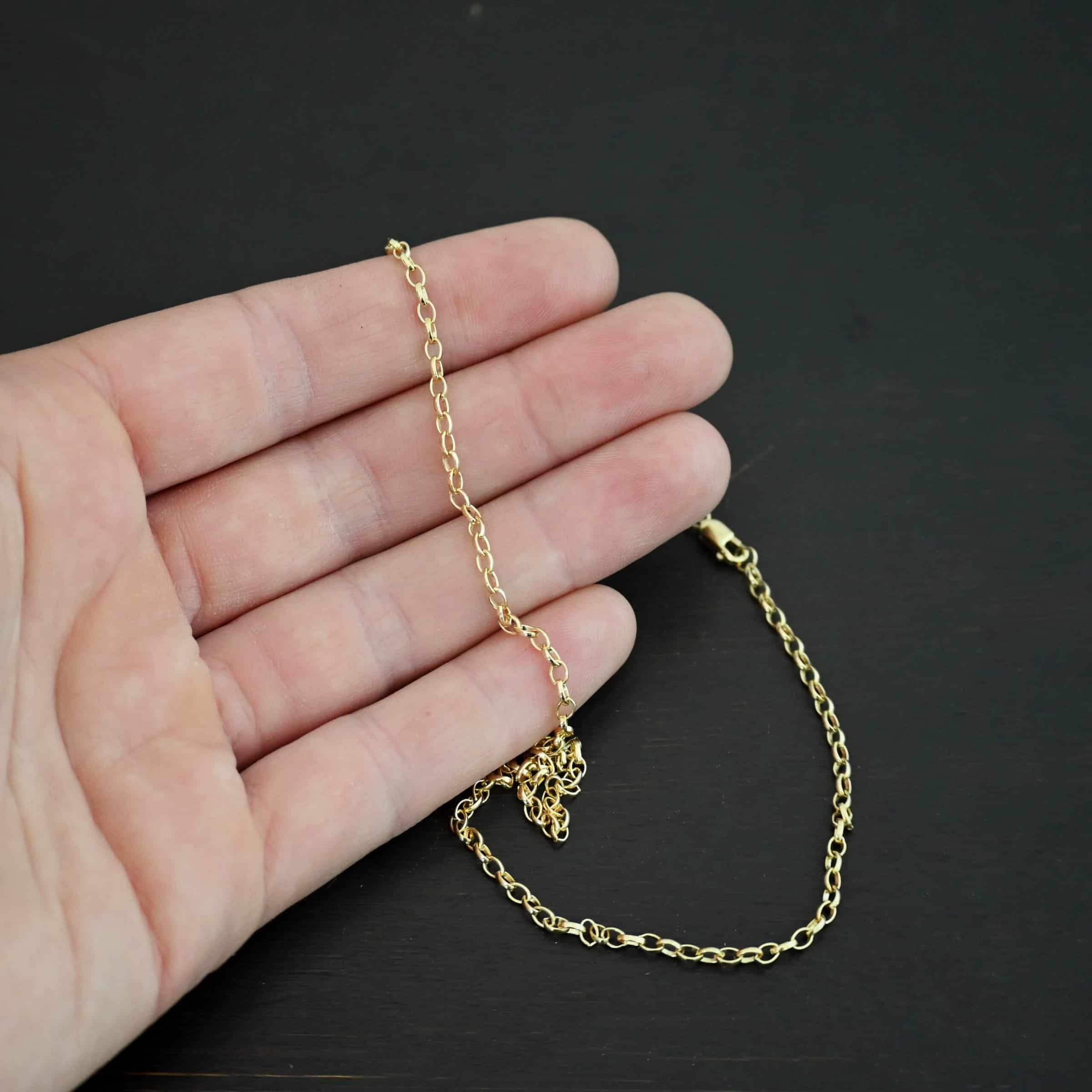 "Ruby Rolo" Chain as shown up close against a dark background. 14K Gold Filled over sterling silver with small circular links connected together. Chain is lying against the fingertips of a hand to demonstrate size.