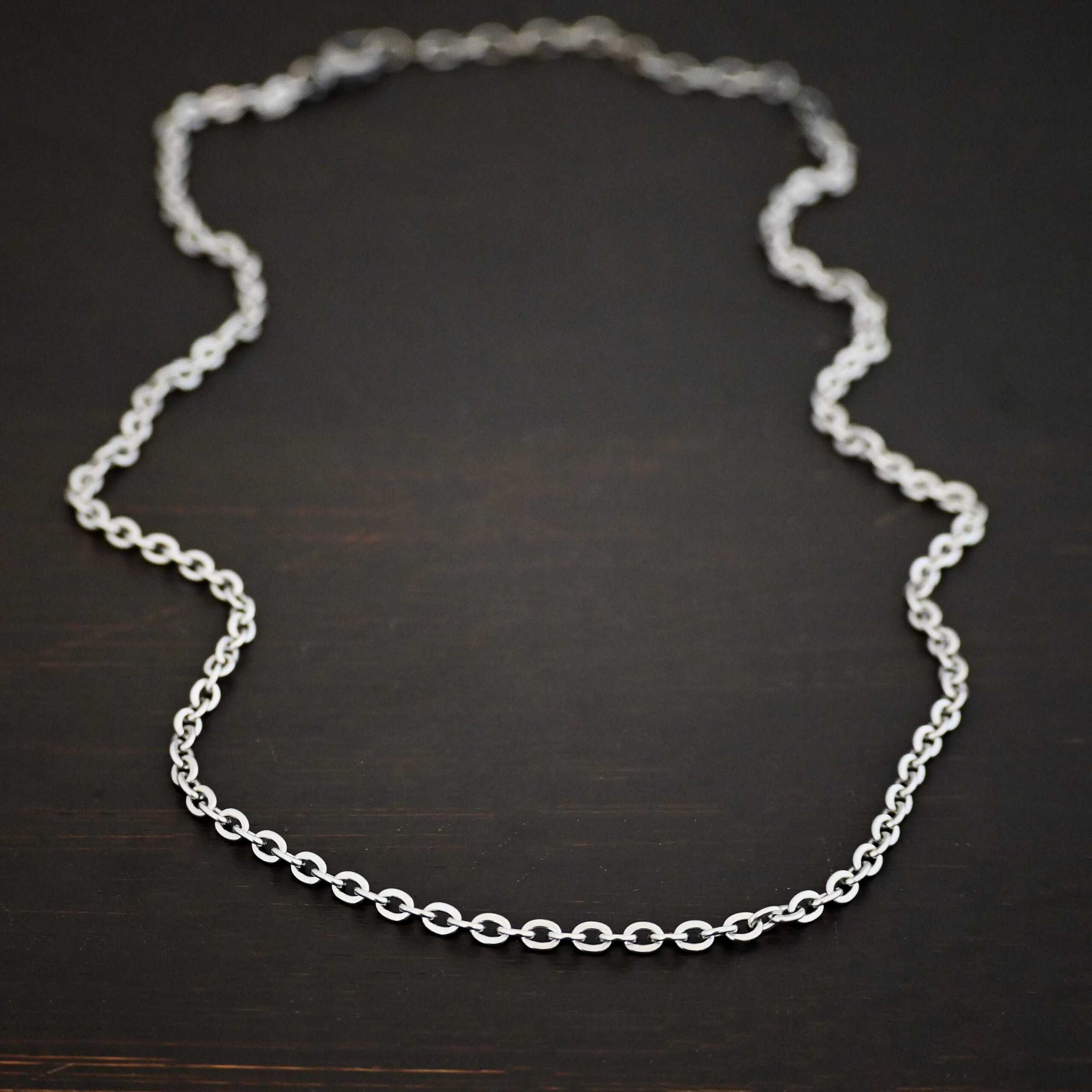 Simon Chain "Simon" Chain as shown up close. 4x3mm links and made out of stainless steel. Chain is laying against a dark background.