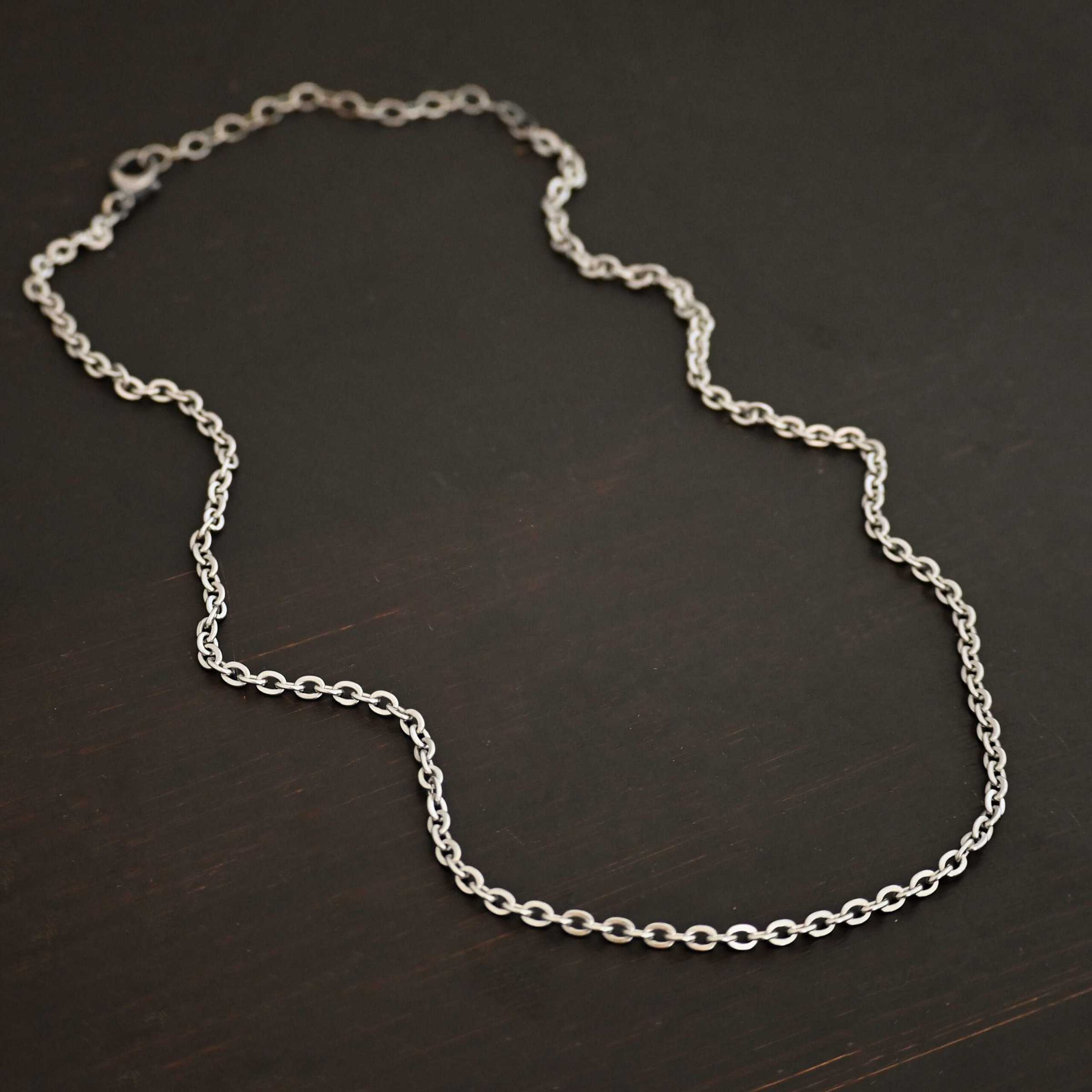 Simon Chain "Simon" Chain as shown up close. 4x3mm links and made out of stainless steel. Chain is laying against a dark background.