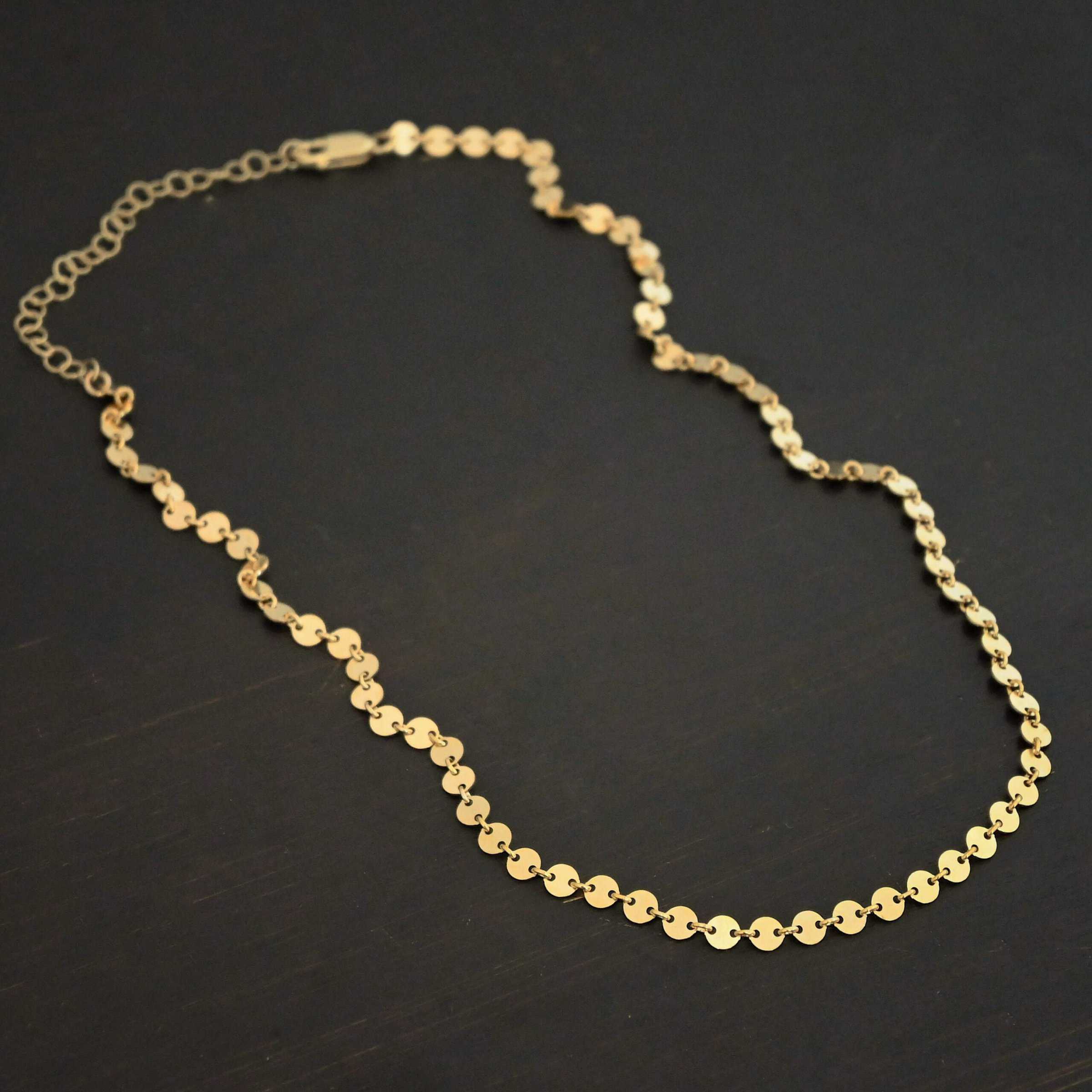 "Gypsy Coin" Chain shown as gold filled. 4mm coin disks connected with jump rings. Each coin has a two holes, one on the left side of the coin and one on the right side. Each hole is filled by a jump ring to connect each coin to one another. Chain is against a dark background.