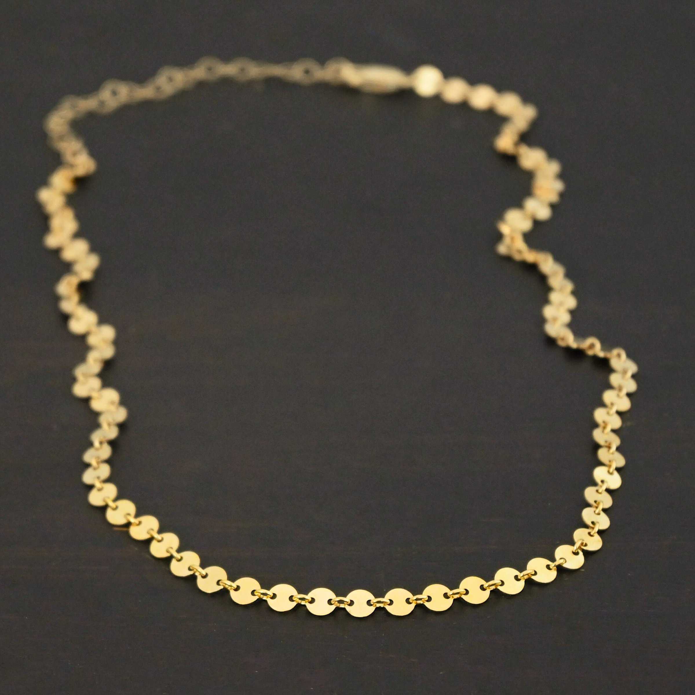"Gypsy Coin" Chain shown as gold filled. 4mm coin disks connected with jump rings. Each coin has a two holes, one on the left side of the coin and one on the right side. Each hole is filled by a jump ring to connect each coin to one another. Chain is against a dark background.
