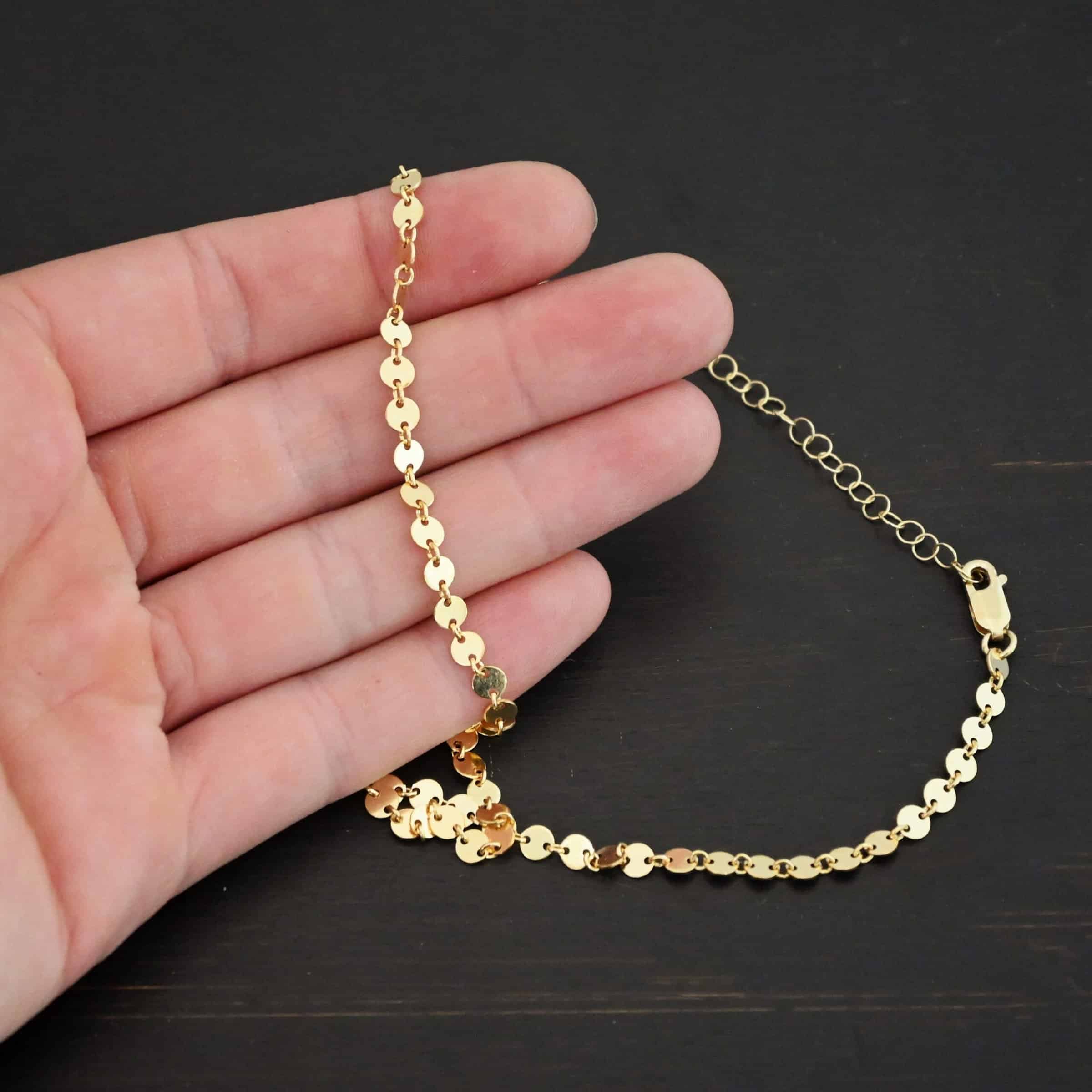 "Gypsy Coin" Chain shown as gold filled. 4mm coin disks connected with jump rings. Each coin has a two holes, one on the left side of the coin and one on the right side. Each hole is filled by a jump ring to connect each coin to one another. Chain is against the fingertips of a hand.