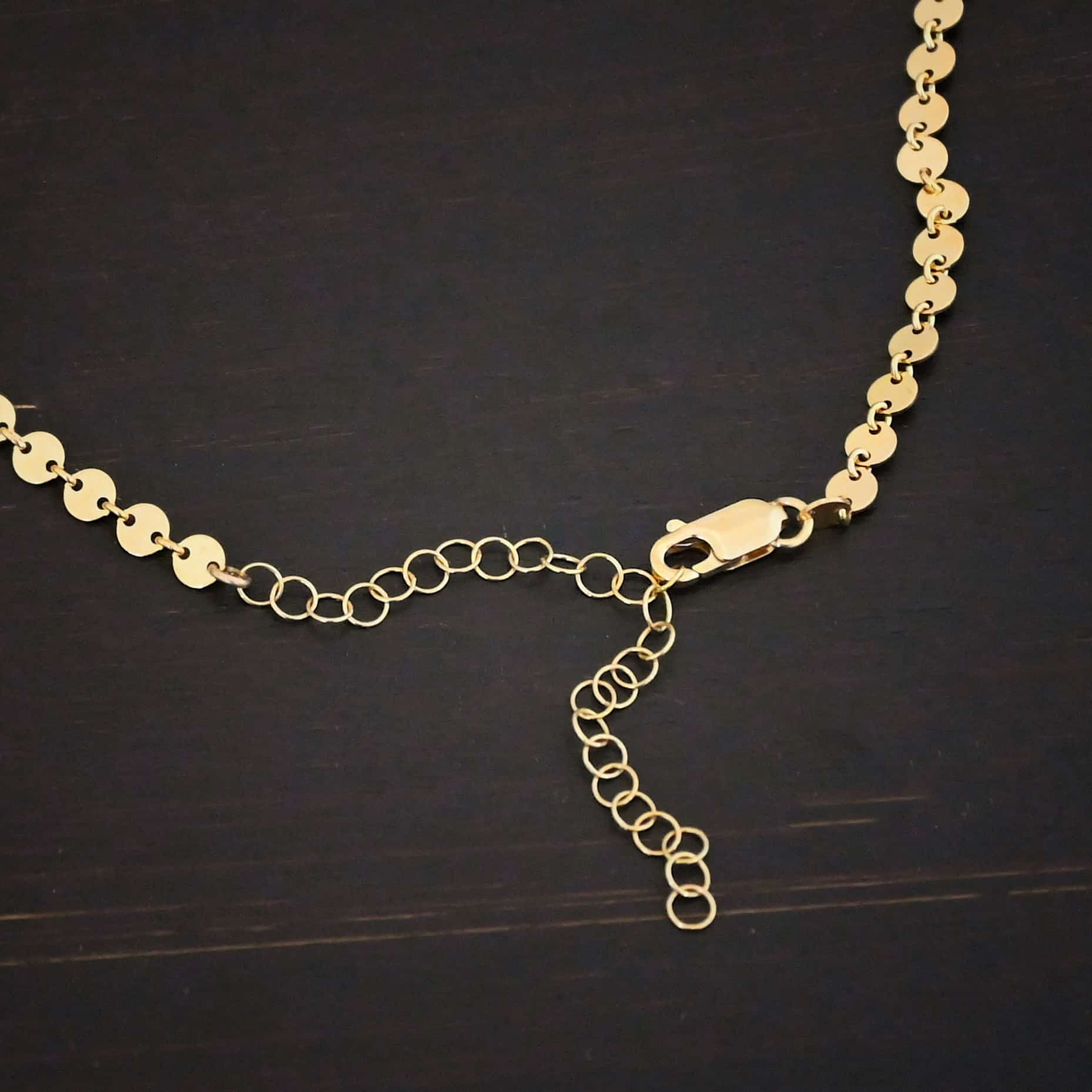"Gypsy Coin" Chain shown as gold filled. 4mm coin disks connected with jump rings. Each coin has a two holes, one on the left side of the coin and one on the right side. Each hole is filled by a jump ring to connect each coin to one another. Chain has a lobster claw clasp and a two inch gold filled extension. Chain is lying against a dark background.
