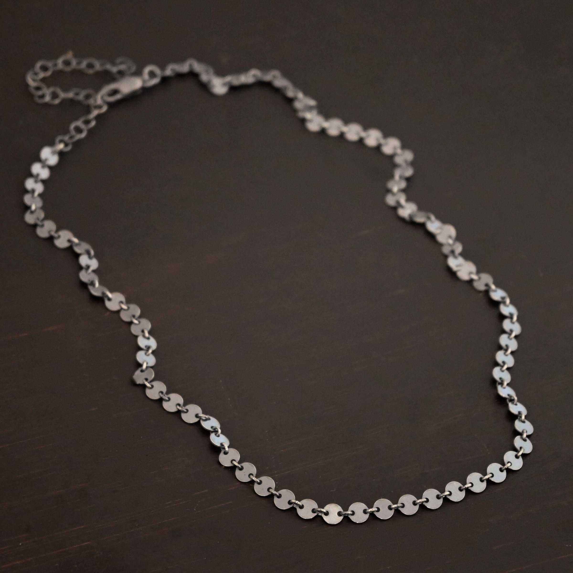"Gypsy Coin" Chain shown as oxidized sterling silver. 4mm coin disks connected with jump rings. Each coin has a two holes, one on the left side of the coin and one on the right side. Each hole is filled by a jump ring to connect each coin to one another. Chain is against a dark background.