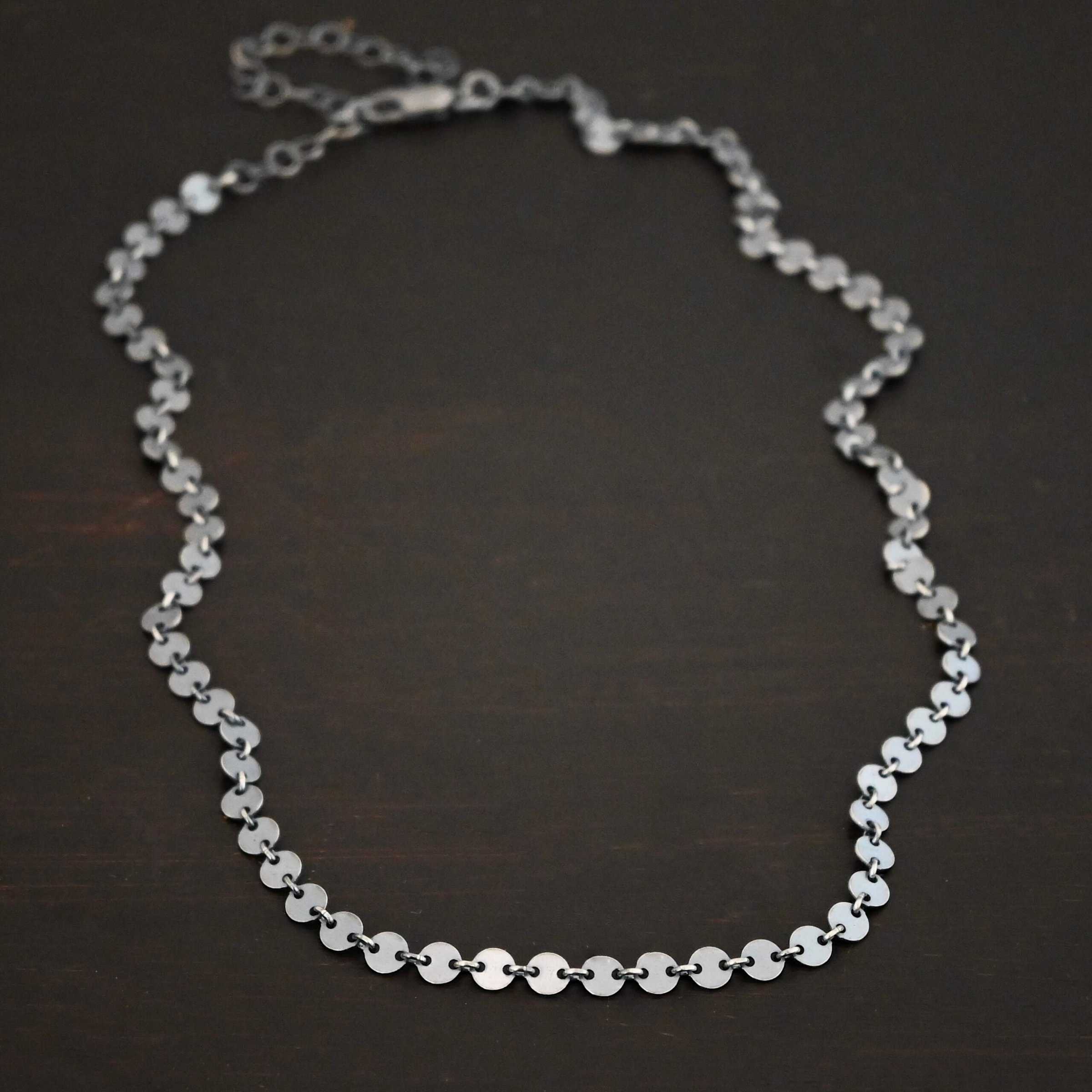 "Gypsy Coin" Chain shown as oxidized sterling silver. 4mm coin disks connected with jump rings. Each coin has a two holes, one on the left side of the coin and one on the right side. Each hole is filled by a jump ring to connect each coin to one another. Chain is against a dark background.