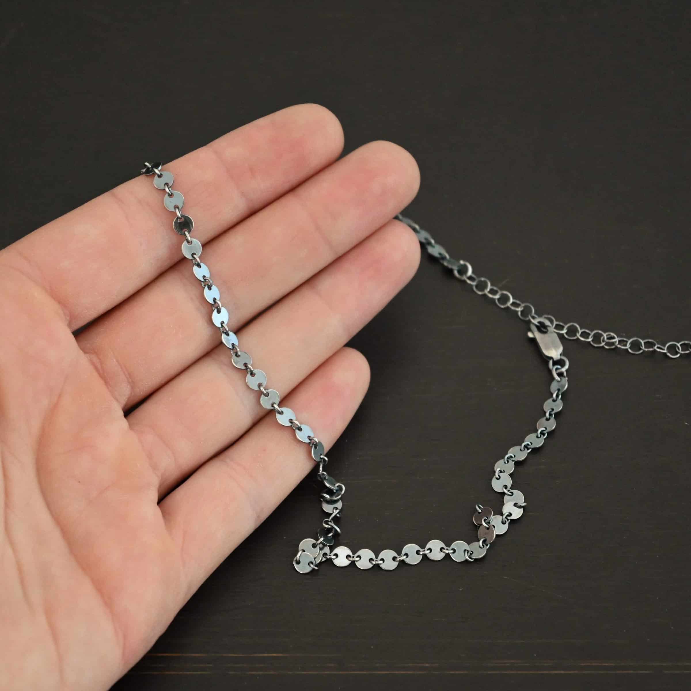 "Gypsy Coin" Chain shown as oxidized sterling silver. 4mm coin disks connected with jump rings. Each coin has a two holes, one on the left side of the coin and one on the right side. Each hole is filled by a jump ring to connect each coin to one another. Chain is lying against the fingertips of a hand.