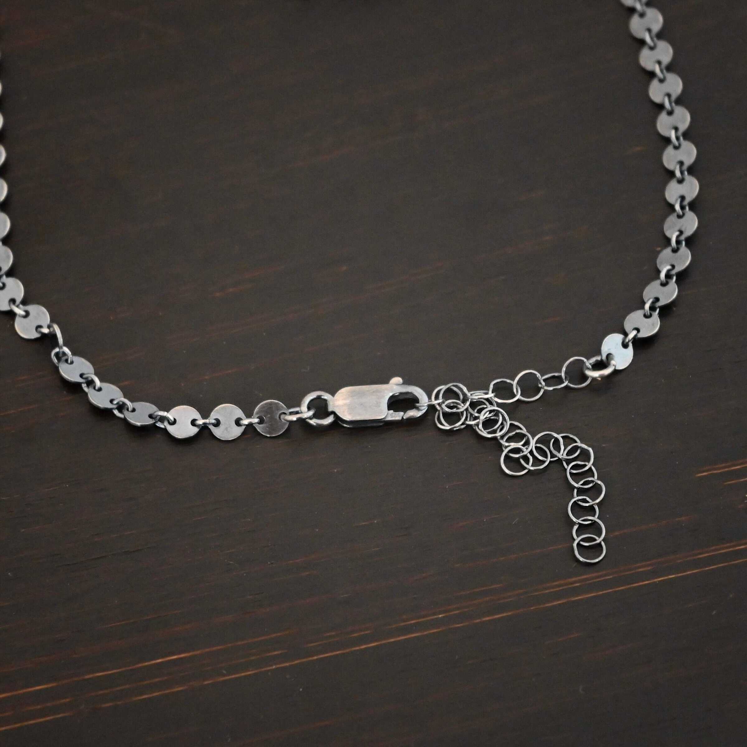 "Gypsy Coin" Chain shown as oxidized sterling silver. 4mm coin disks connected with jump rings. Each coin has a two holes, one on the left side of the coin and one on the right side. Each hole is filled by a jump ring to connect each coin to one another. Chain has a lobster claw clasp and a two inch sterling silver extension. Chain is lying against a dark background.