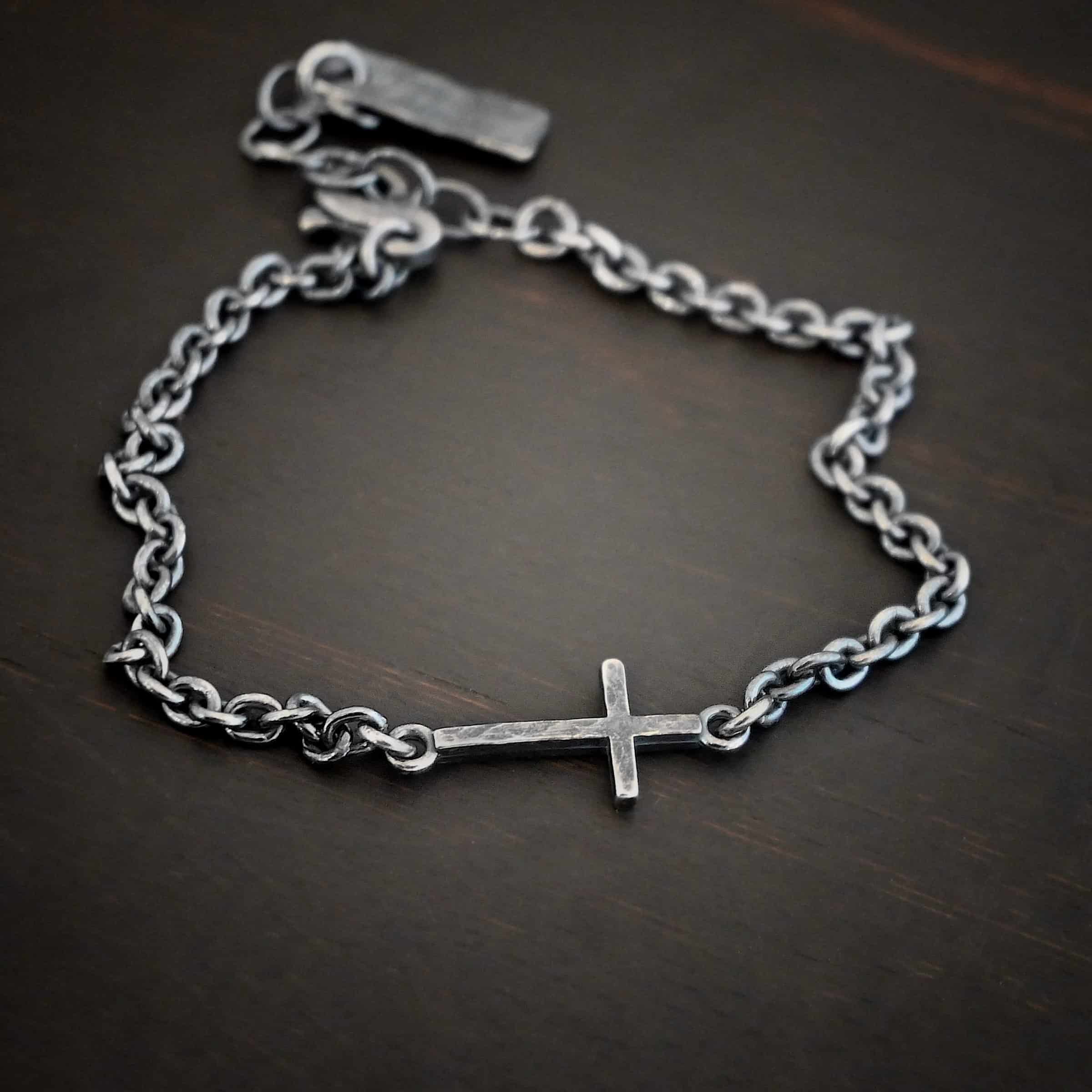 Photo of the "Creator" Bracelet. There is a cross set horizontally with a loop on each end. Each end of the cross is connected to each side of the chain in the center of the bracelet. The bracelet is oxidized, and lying against a dark background.