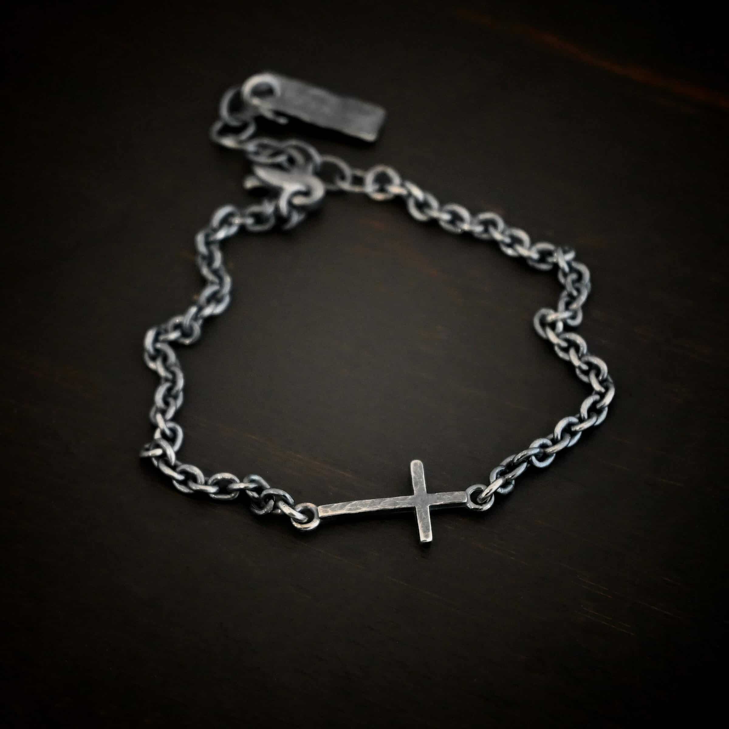 Photo of the "Creator" Bracelet. There is a cross set horizontally with a loop on each end. Each end of the cross is connected to each side of the chain in the center of the bracelet. The bracelet is oxidized, and lying against a dark background.