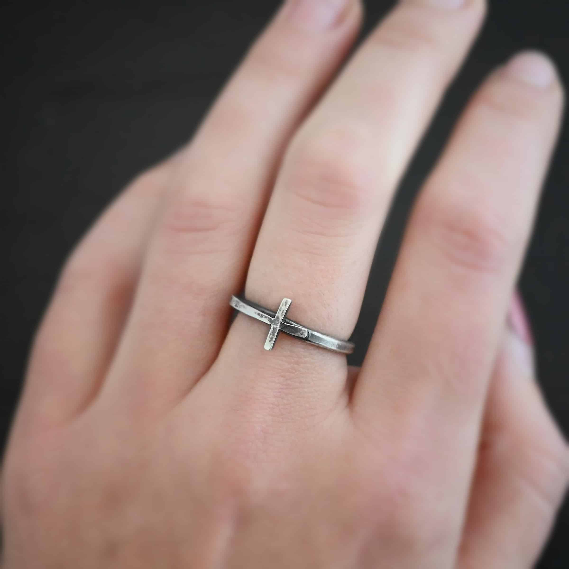 Photo of a creator ring. Ring is made out of sterling silver. The ring band is thin and made of silver. There is a cross that is in the center of the ring band and set sideways. The top of bottom of the cross are soldered onto the ring band. The top side of the cross is sticking into the air and the bottom side of the cross is set on the table. Ring is worn by a model up close.