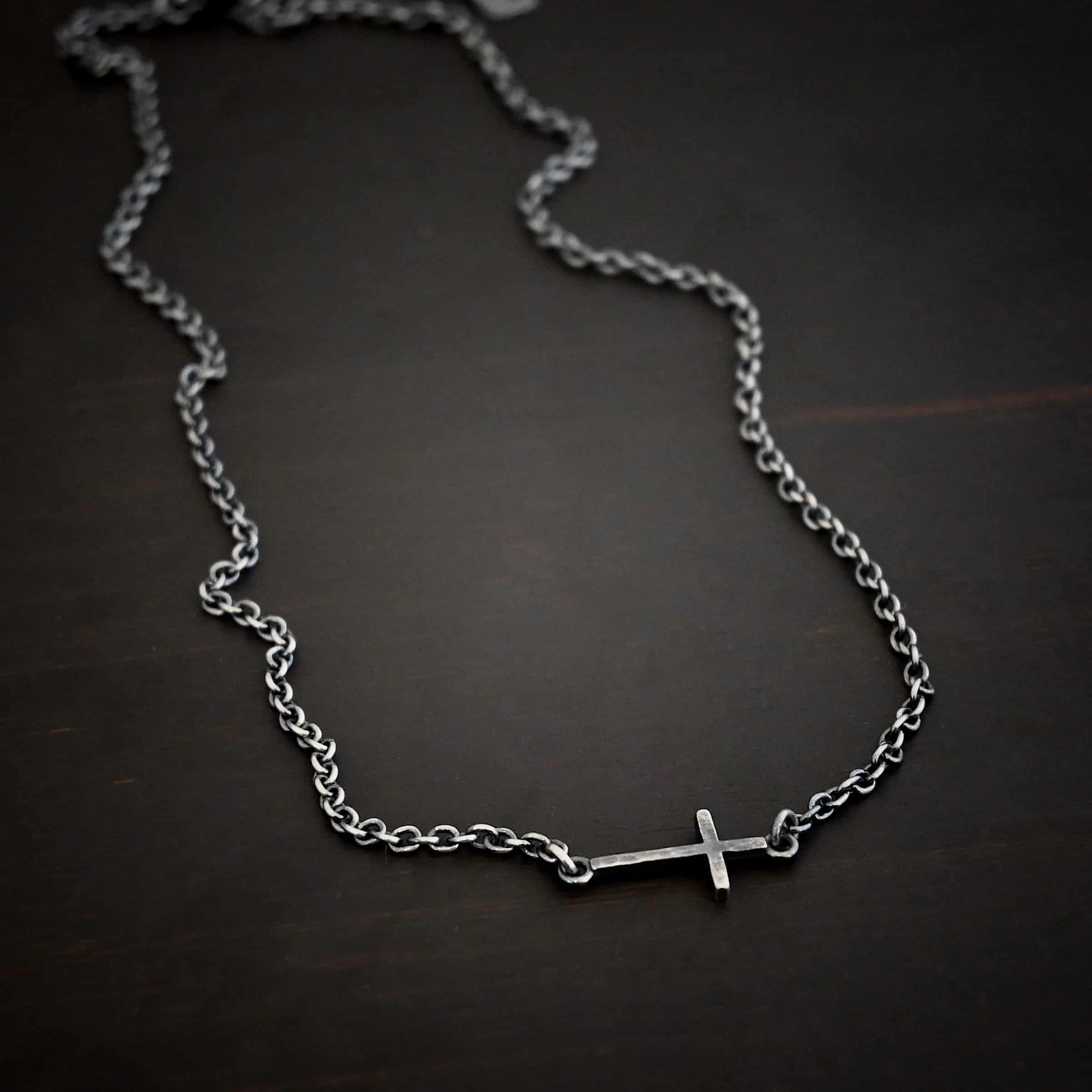 Creator Necklace Photo of the "Creator" Necklace. Made out of sterling silver. There is a small, but thick hammered silver cross hung vertically and attached on both ends to the chain of the necklace. Each end of the cross has a loop on it to attach each end to the chain. Necklace is oxidized and lying against a dark background.