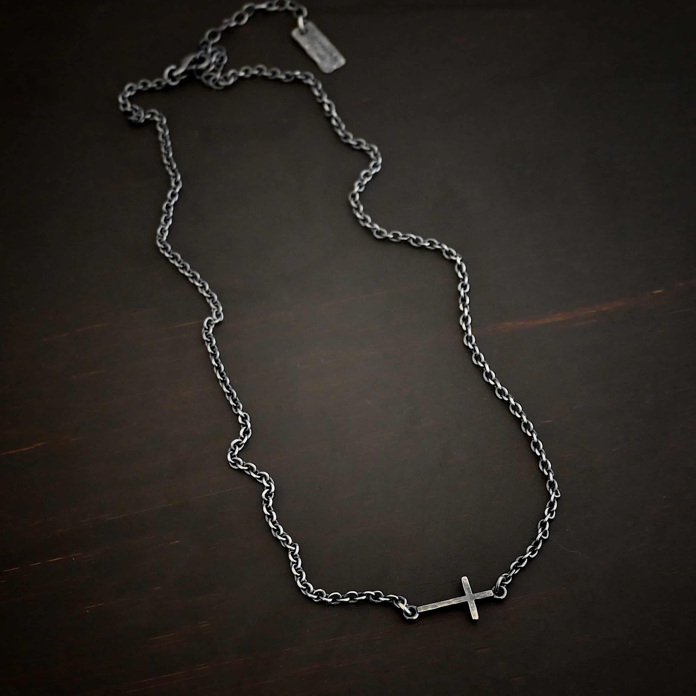 Creator Necklace Photo of the "Creator" Necklace. Made out of sterling silver. There is a small, but thick hammered silver cross hung vertically and attached on both ends to the chain of the necklace. Each end of the cross has a loop on it to attach each end to the chain. Necklace is oxidized and lying against a dark background.