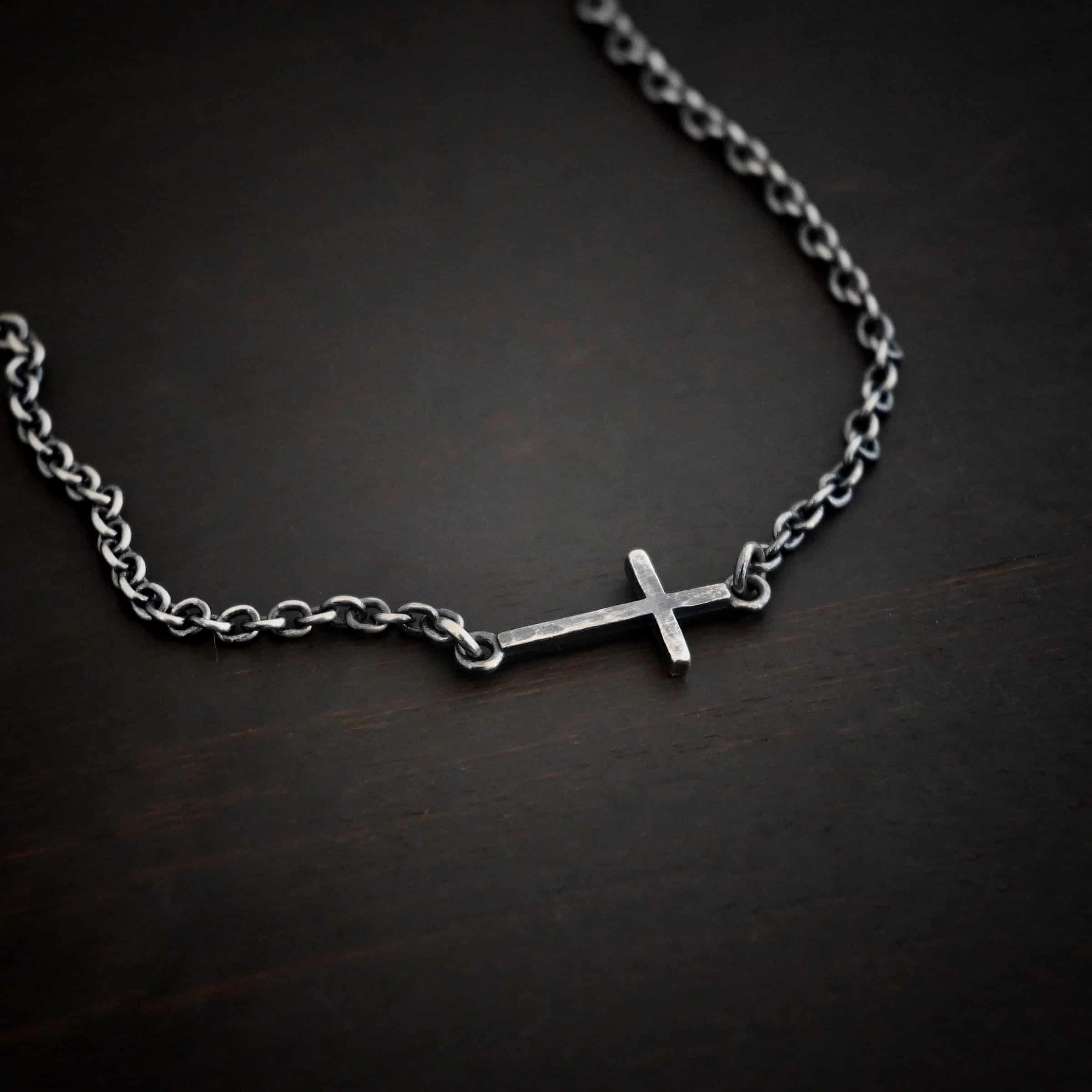 Creator Necklace Photo of the "Creator" Necklace. Made out of sterling silver. There is a small, but thick hammered silver cross hung vertically and attached on both ends to the chain of the necklace. Each end of the cross has a loop on it to attach each end to the chain. Necklace is oxidized and lying against a dark background.