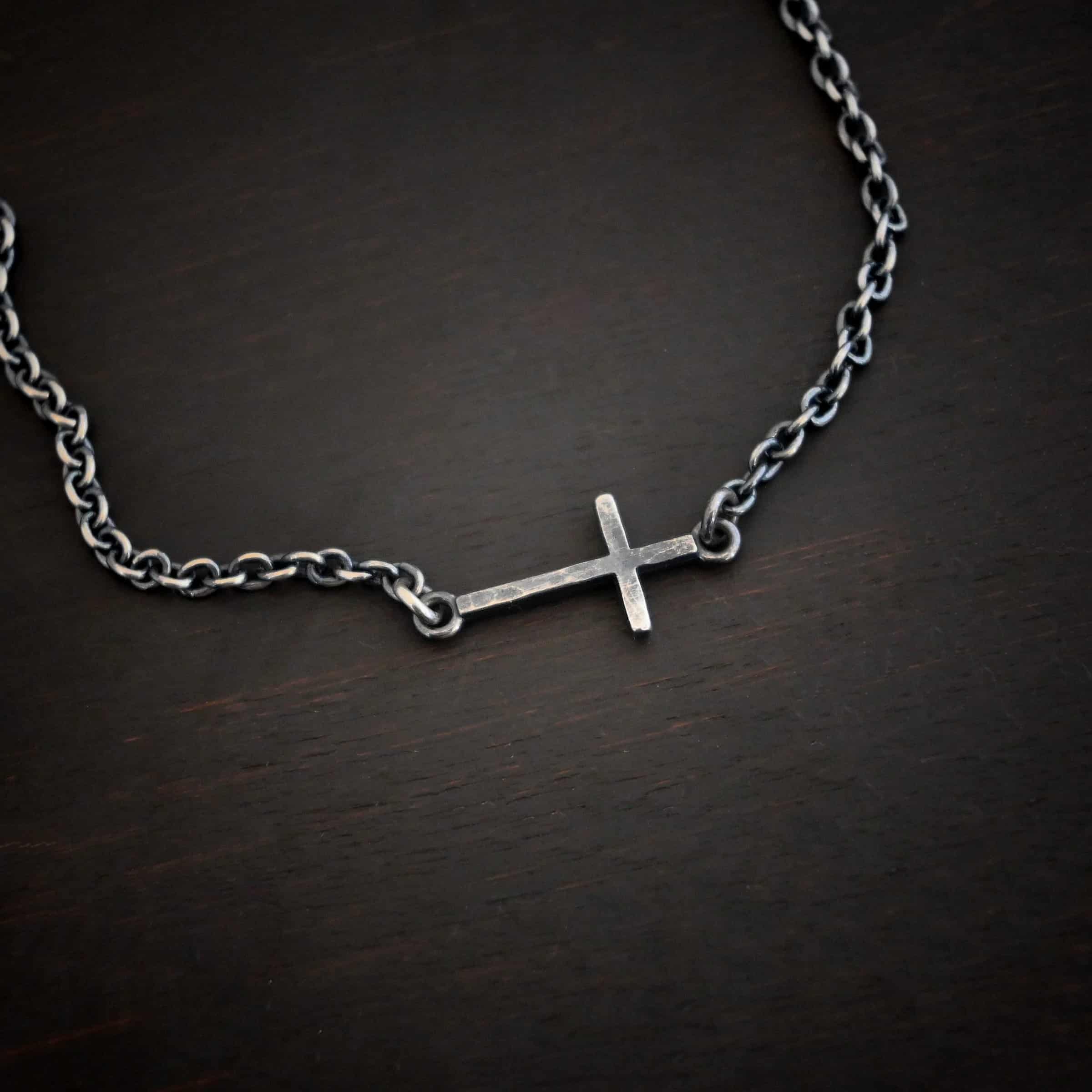 Creator Necklace Photo of the "Creator" Necklace. Made out of sterling silver. There is a small, but thick hammered silver cross hung vertically and attached on both ends to the chain of the necklace. Each end of the cross has a loop on it to attach each end to the chain. Necklace is oxidized and lying against a dark background.