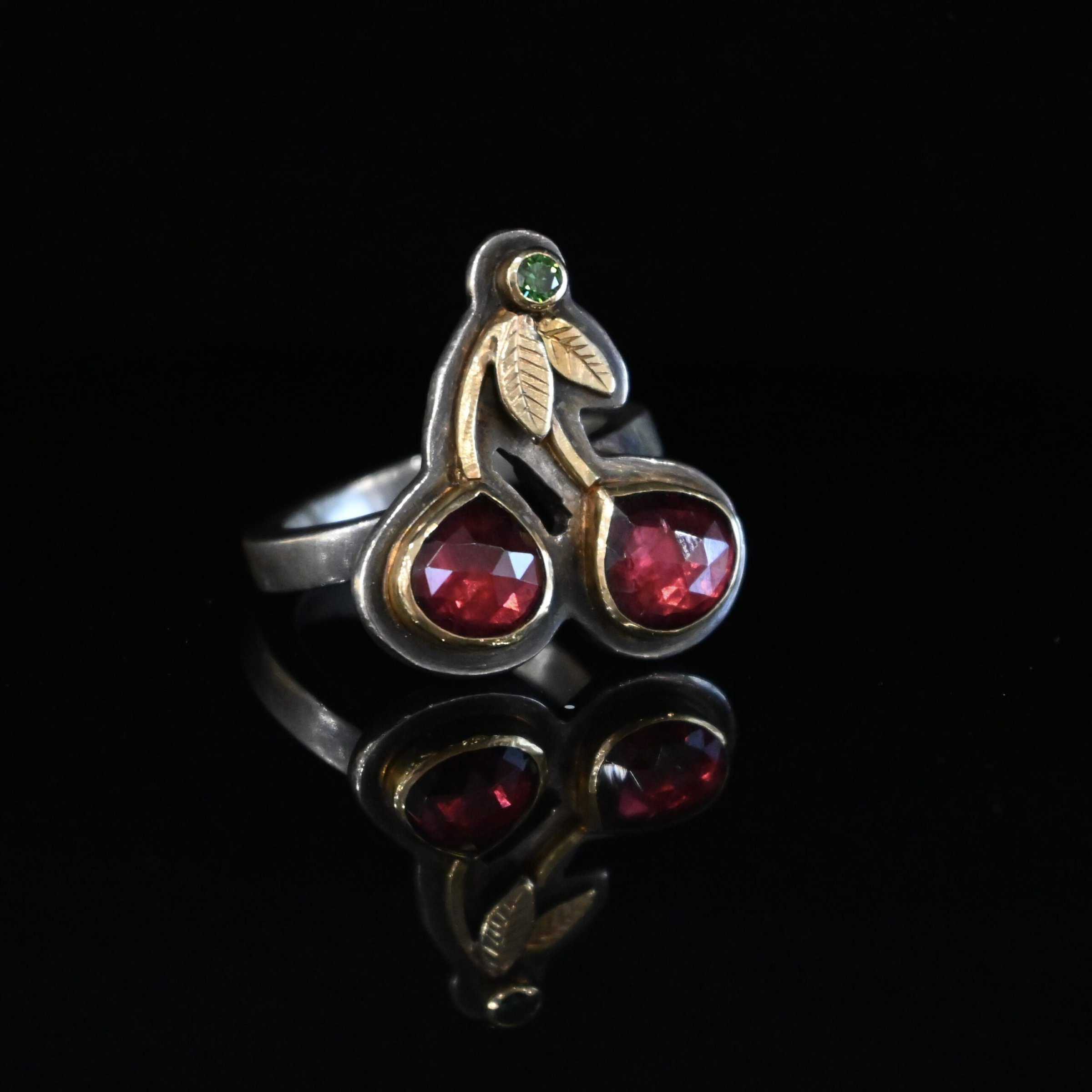 Door County Cherry Ring Close up of the "Door County Cherry" Ring. Made out of sterling silver, gold, garnets, and green diamond. There are two slightly teardrop shaped garnets that make up the "Cherry" part of the ring. They are set in gold, and the gold leads up to the small circular green diamond as a part of the "stem" of the cherries. There are two gold leaves on the ring as well. Ring is set against a dark background.