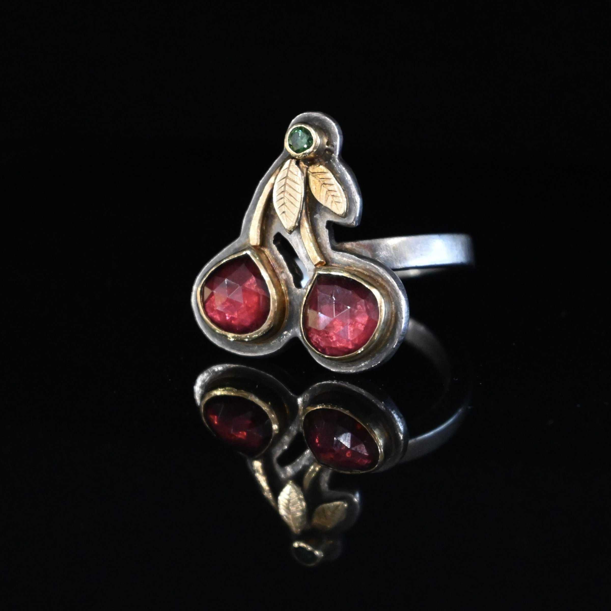 Door County Cherry Ring Close up of the "Door County Cherry" Ring. Made out of sterling silver, gold, garnets, and green diamond. There are two slightly teardrop shaped garnets that make up the "Cherry" part of the ring. They are set in gold, and the gold leads up to the small circular green diamond as a part of the "stem" of the cherries. There are two gold leaves on the ring as well. Ring is set against a dark background.
