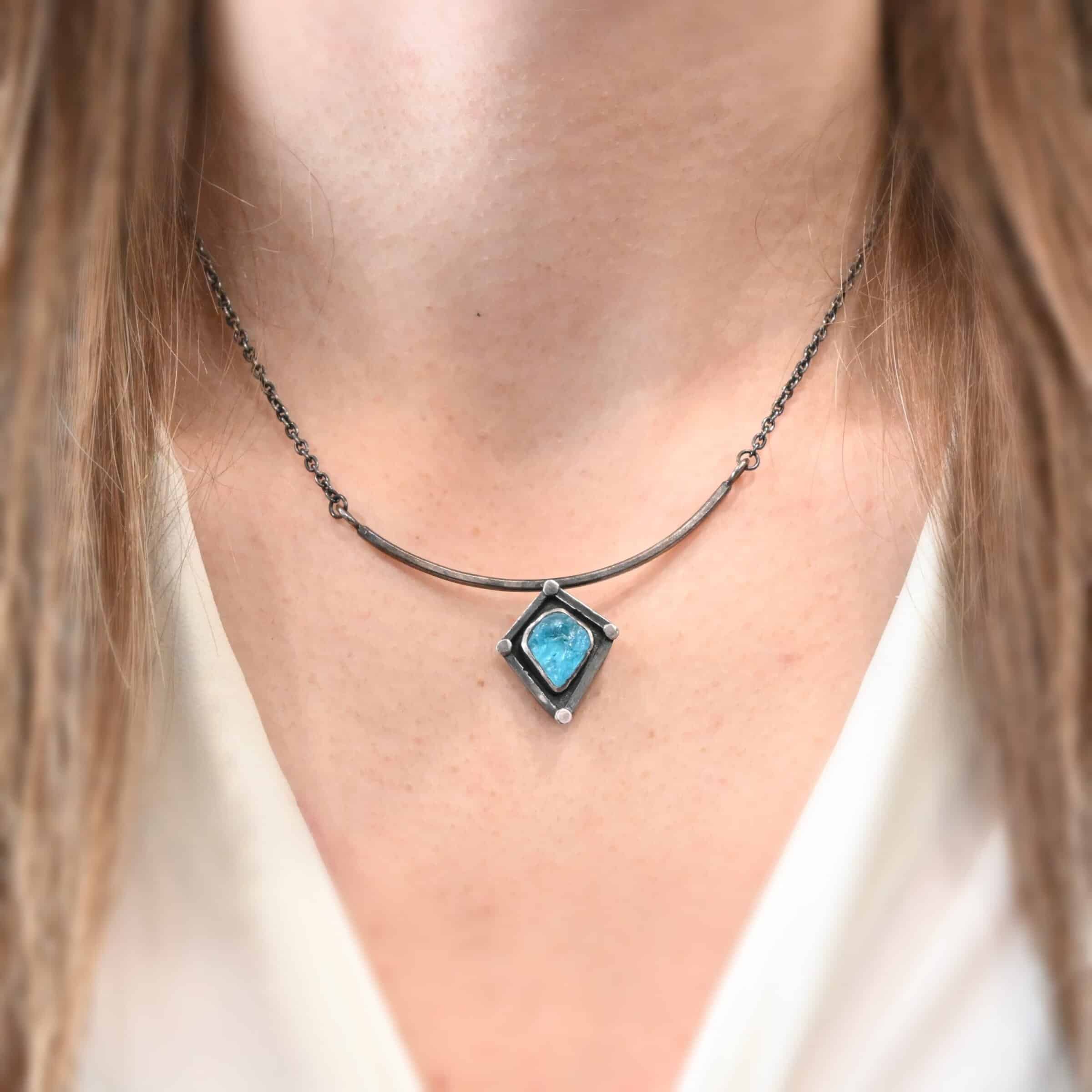 Elsa Necklace "Elsa" Necklace. There is a rough cut apatite in the center of a diamond shaped piece of silver. There is a dot of silver on each point of the silver diamond. The silver diamond is soldered to a curved bar of silver.