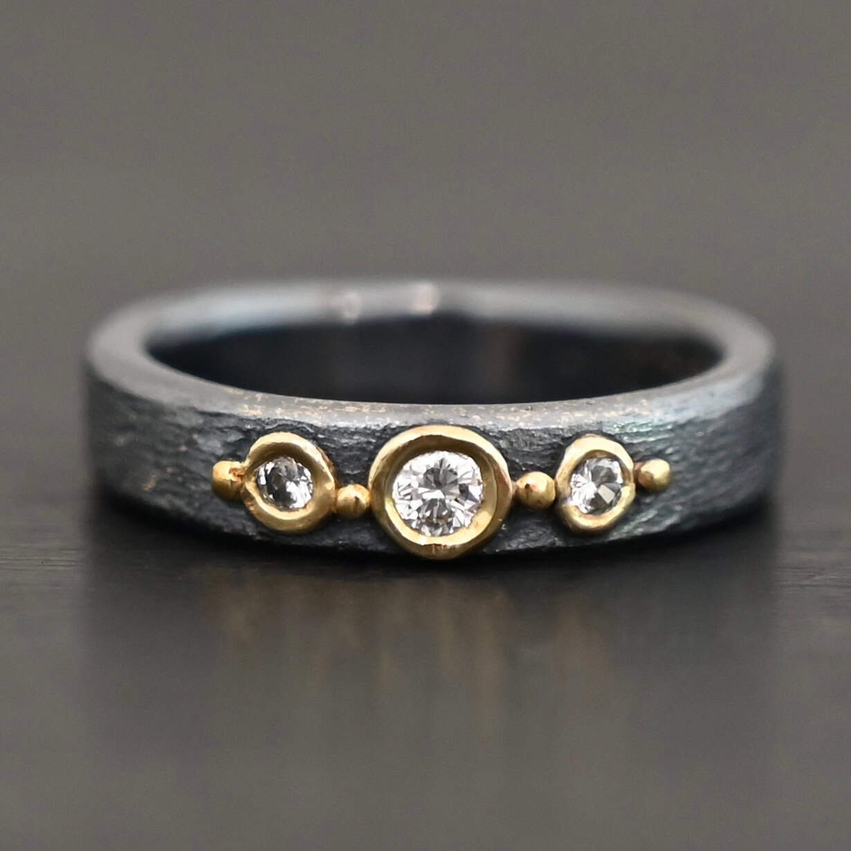 Photo of the "Adoration" Ring. Made out of sterling silver, gold, and diamonds. There are three diamonds that go straight across the center of the ring. Each one is set in gold, and each diamond is separated by a ball of gold. There are four balls of gold in total. The rest of the ring is oxidized and has a textured look to it.