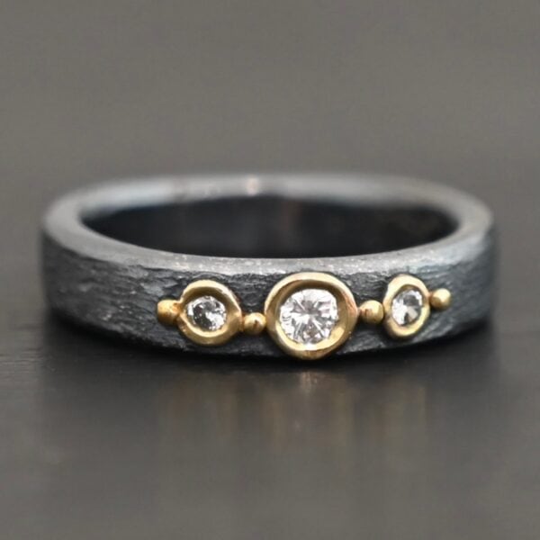 Photo of the "Adoration" Ring. Made out of sterling silver, gold, and diamonds. There are three diamonds that go straight across the center of the ring. Each one is set in gold, and each diamond is separated by a ball of gold. There are four balls of gold in total. The rest of the ring is oxidized and has a textured look to it.